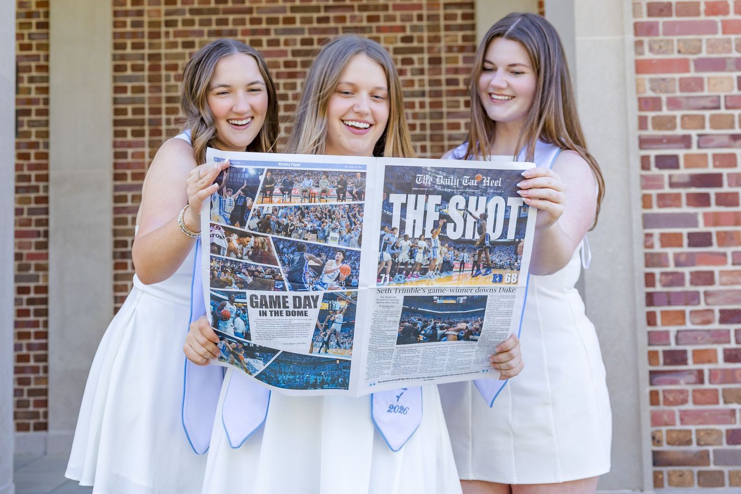 Three graduates in white dresses holding a campus newspaper called The Shot, smiling outdoors near brick building.