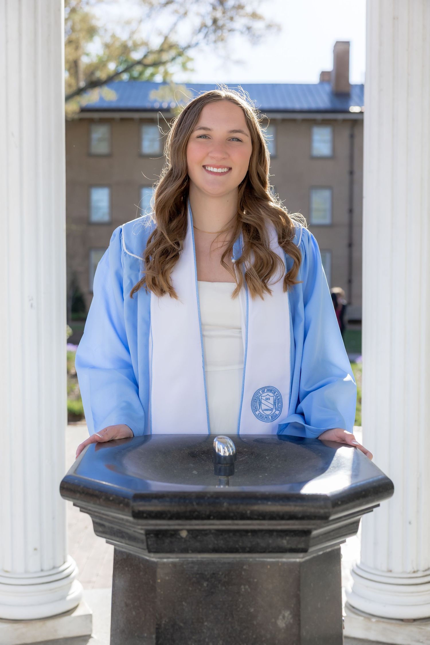 Smiling young woman in light blue graduation gown and white stole stands at a stone podium between white columns.