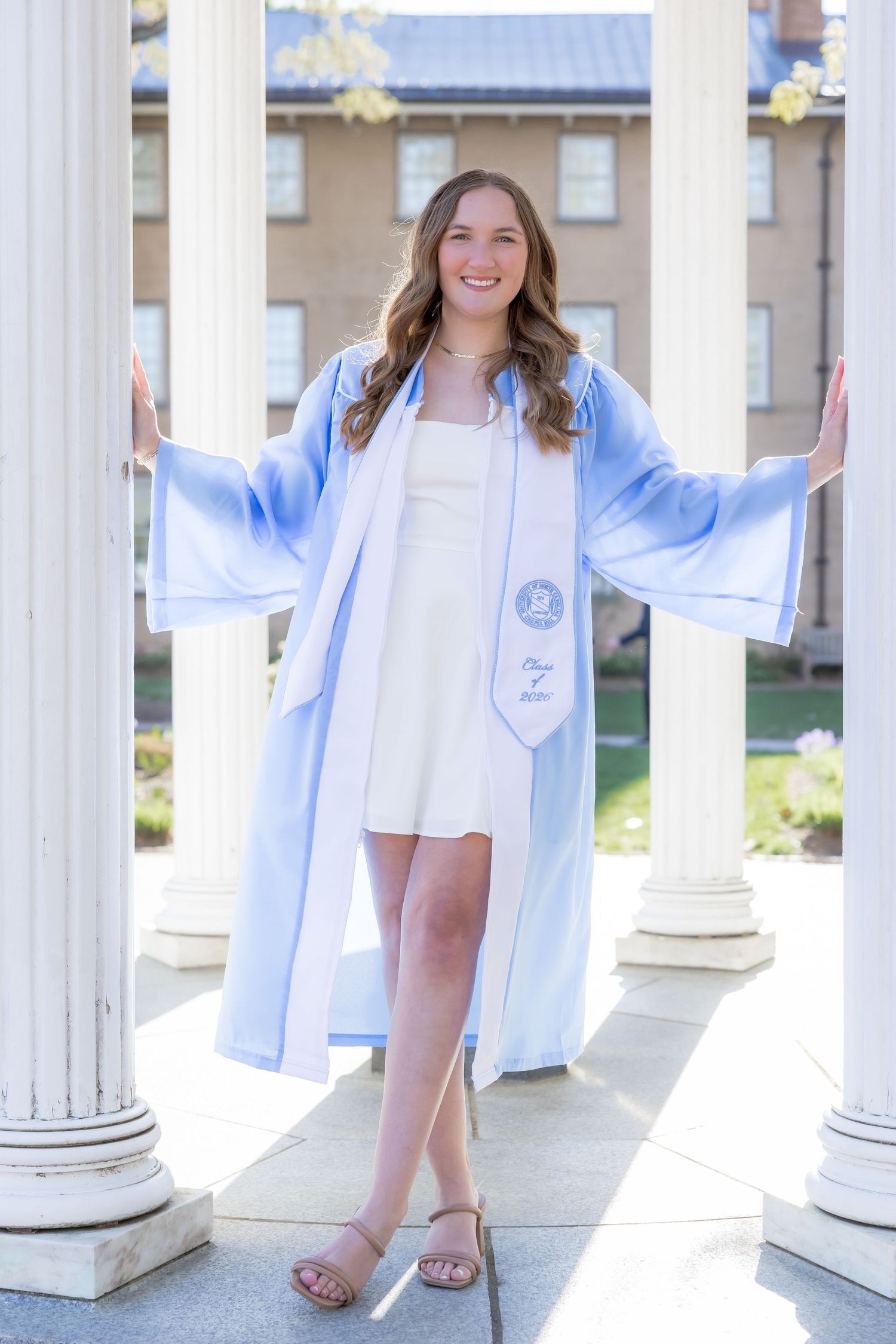 Smiling graduate in light blue cap and gown poses between white columns outdoors on college campus.