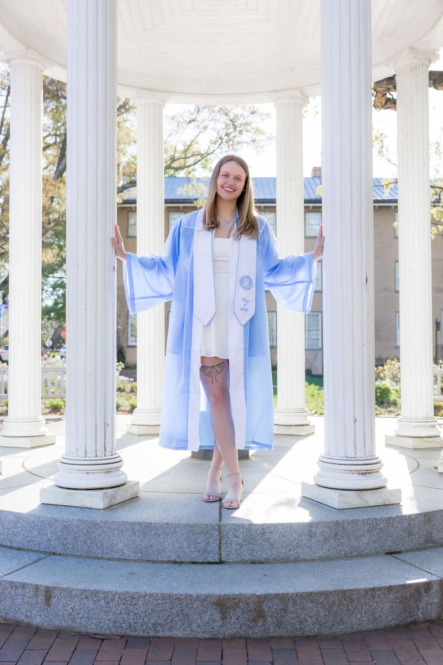 Smiling graduate in light blue cap and gown poses between white columns at UNC Chapel Hill's Old Well.