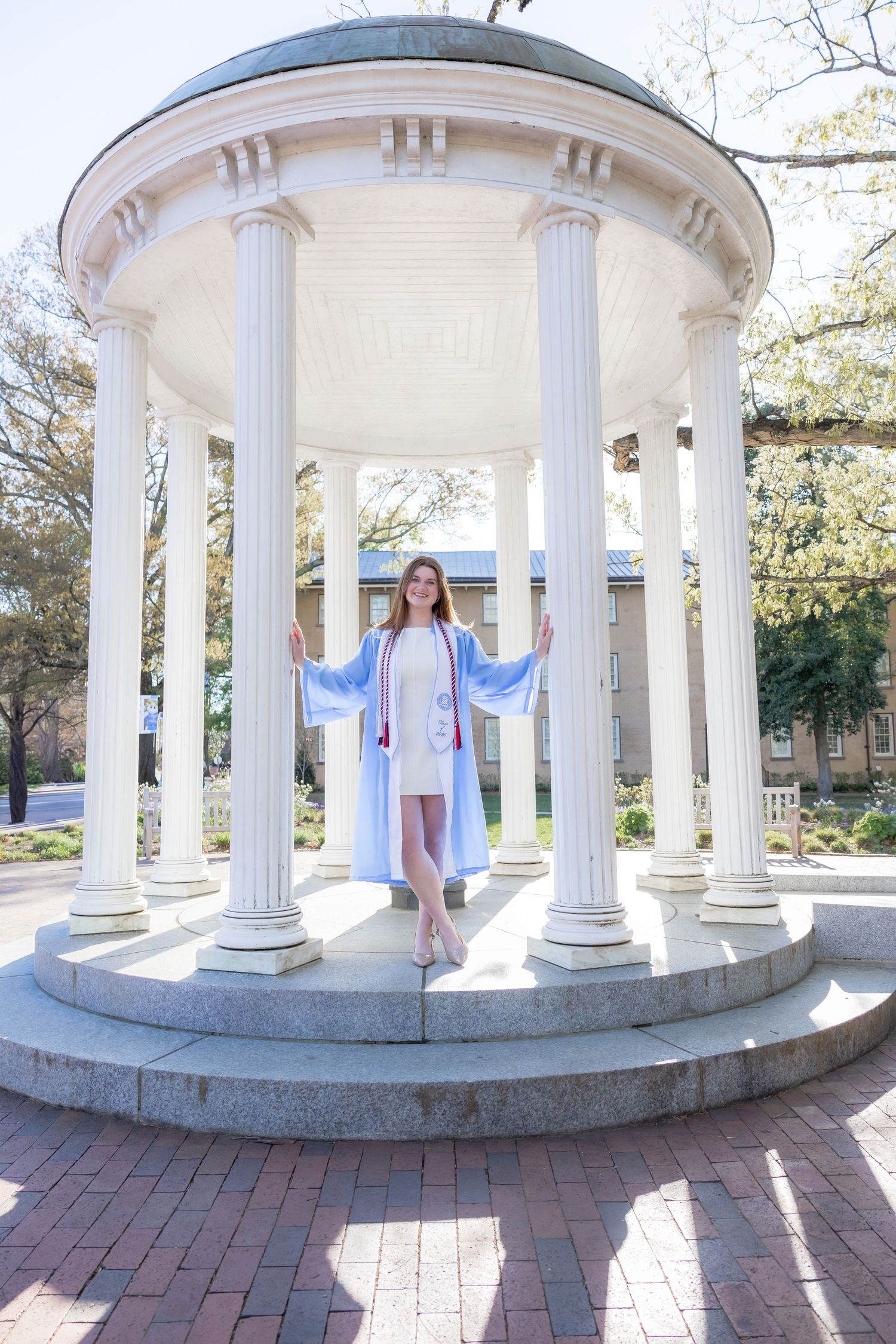 Graduate in light blue cap and gown poses proudly at the Old Well rotunda on UNC Chapel Hill campus.
