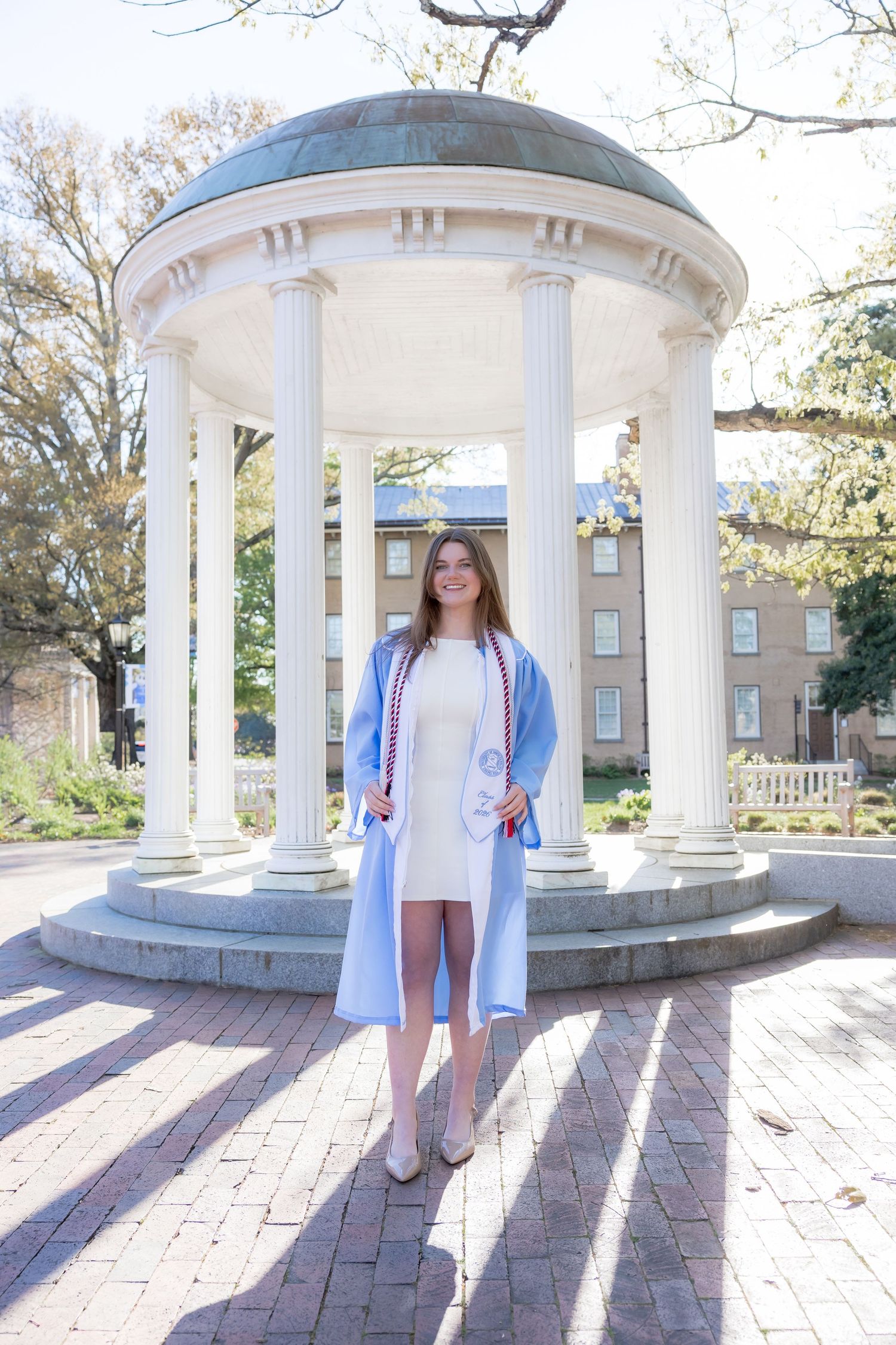A graduate poses in a light blue gown at UNC Chapel Hill's Old Well landmark on a sunny day.