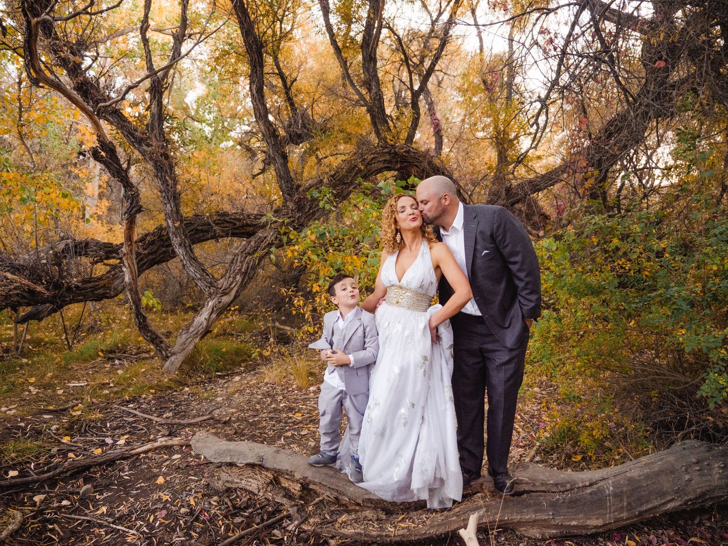 A couple in wedding attire with a young boy pose among autumn trees in a romantic outdoor fall forest setting.