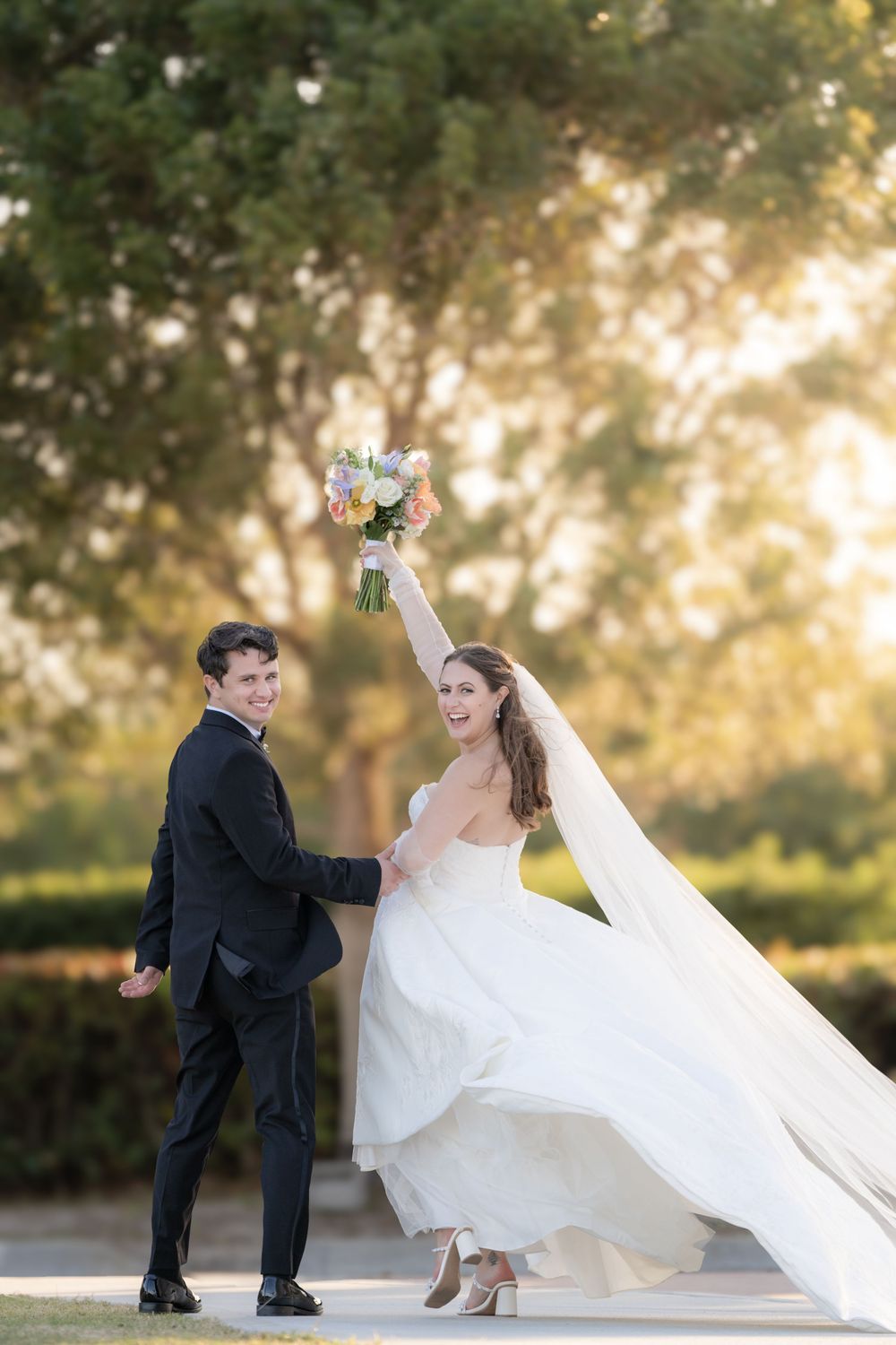 Joyful bride raises colorful bouquet while walking with groom in black suit at golden hour outdoor wedding.