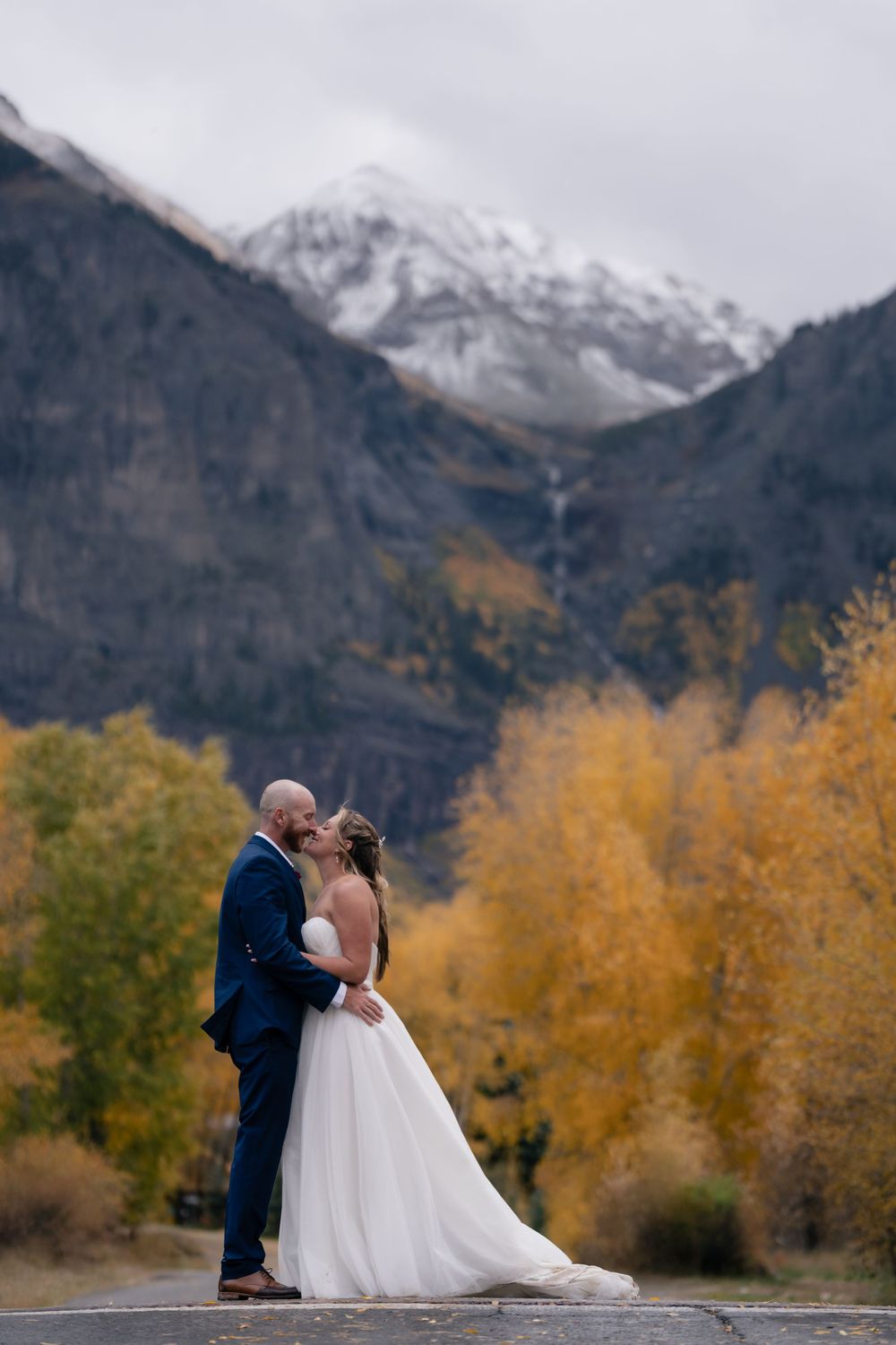 Bride and groom kissing in front of snow-capped mountains and golden autumn trees in a romantic outdoor wedding photo.