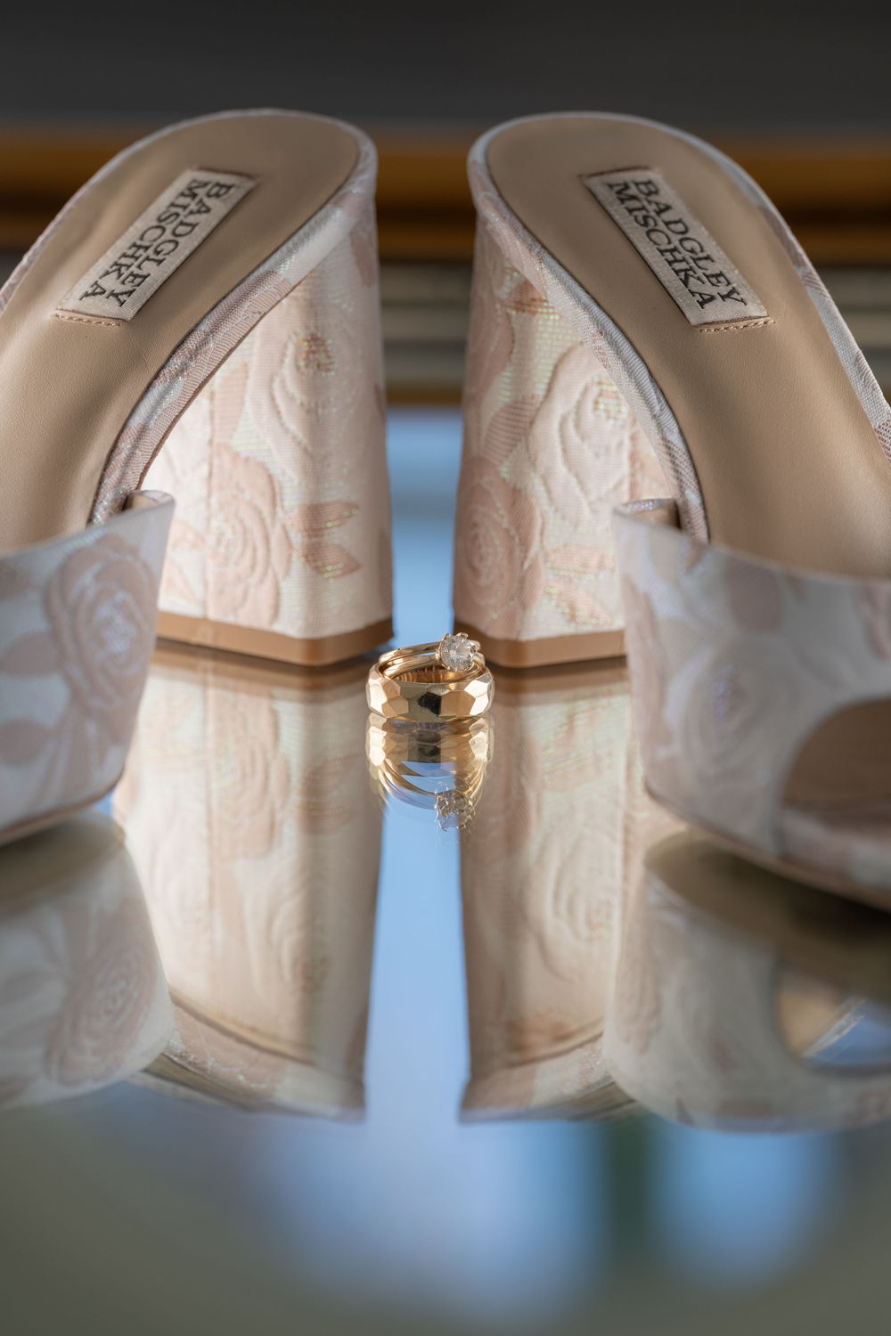 Badgley Mischka bridal wedge heels with floral pattern displayed with gold wedding rings on reflective surface.