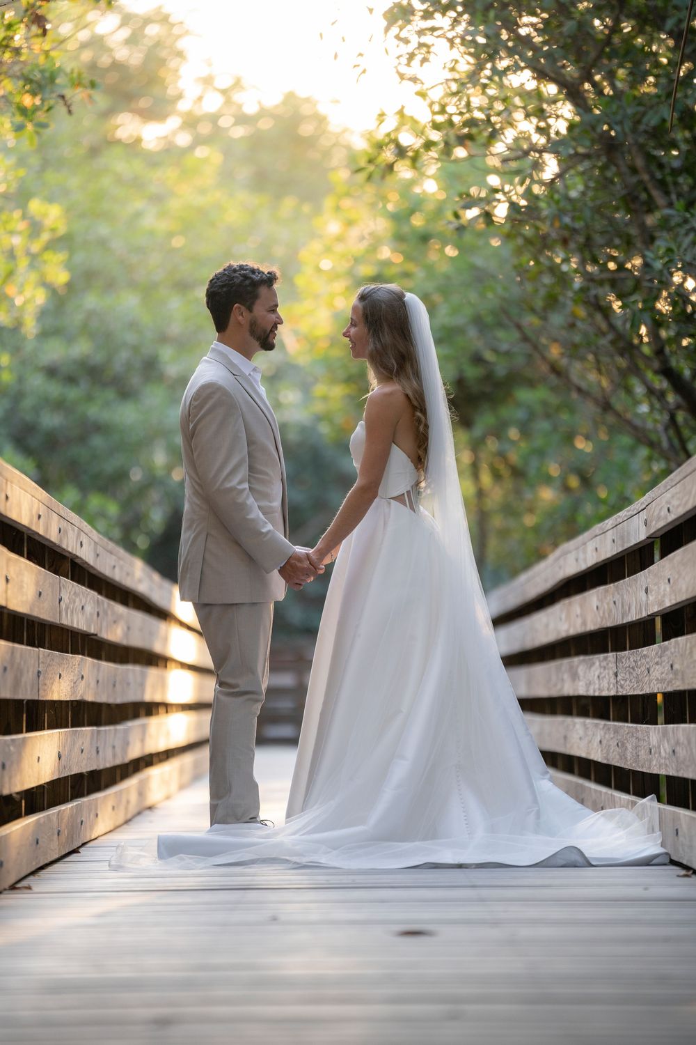 Bride and groom holding hands on a wooden bridge surrounded by lush green trees in golden hour light.