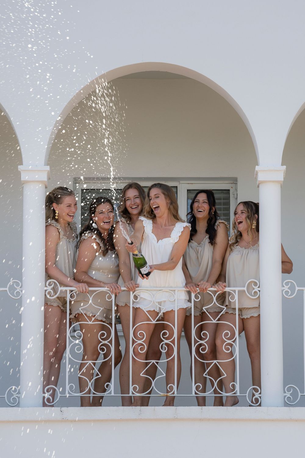 Women celebrating on a balcony with champagne popping, laughing joyfully in elegant attire at a bachelorette party.