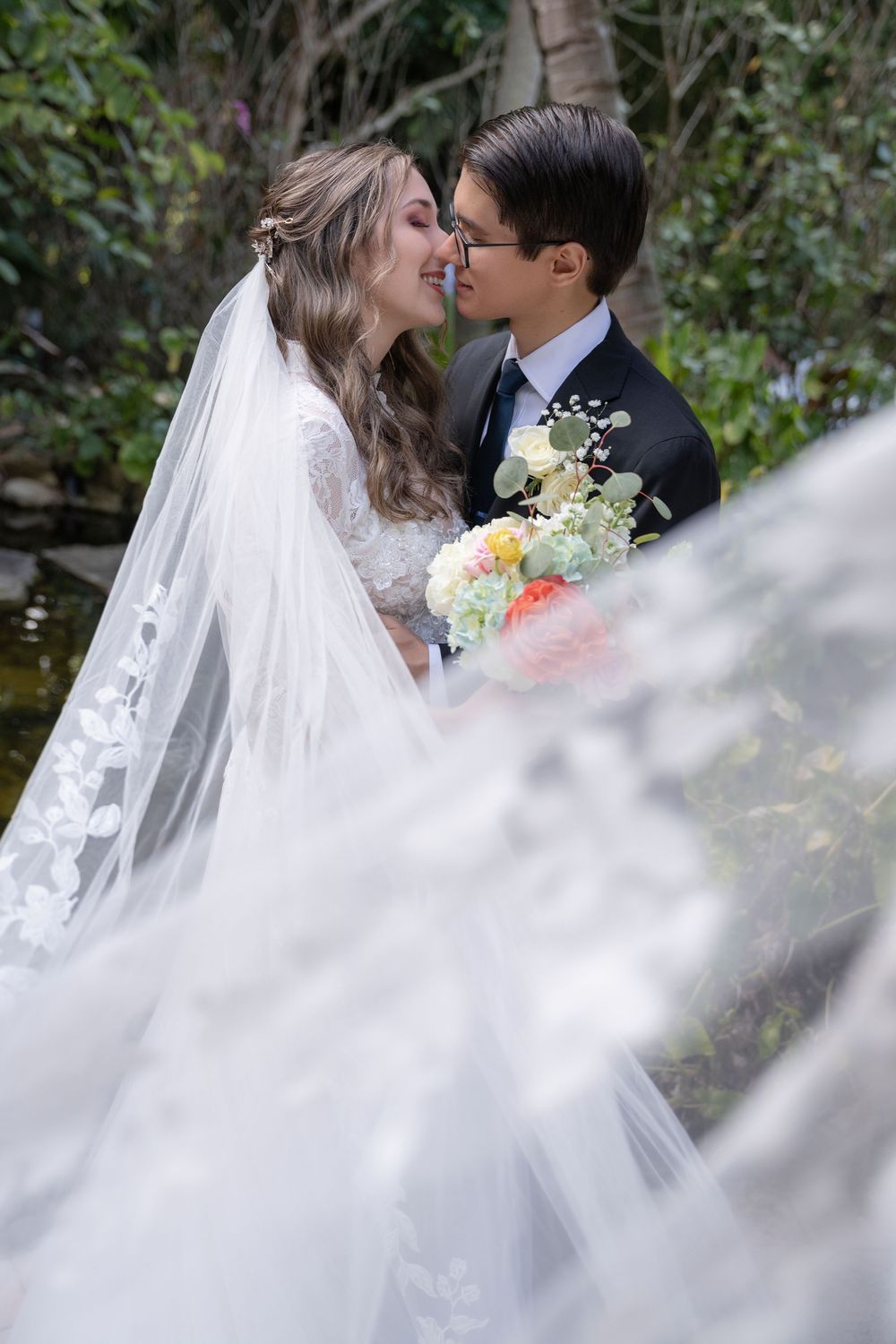Bride and groom share a romantic moment outdoors, with flowing white veil and colorful bouquet amid lush greenery.