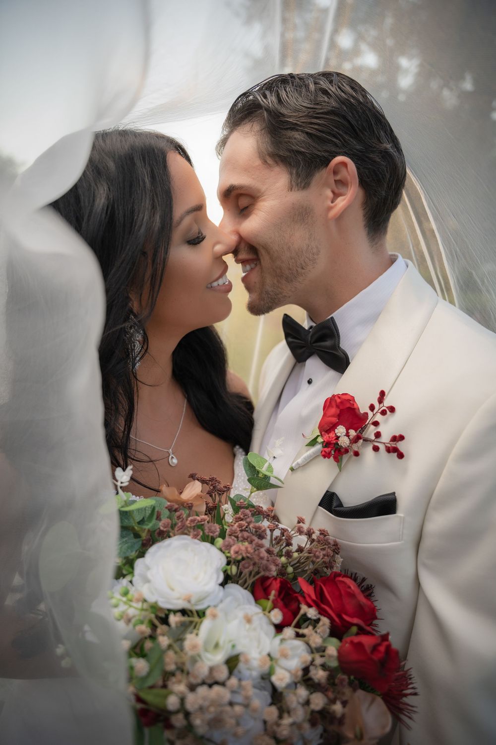 Bride and groom nearly kissing under veil, groom in white tuxedo with red rose boutonniere, bride holding bouquet.