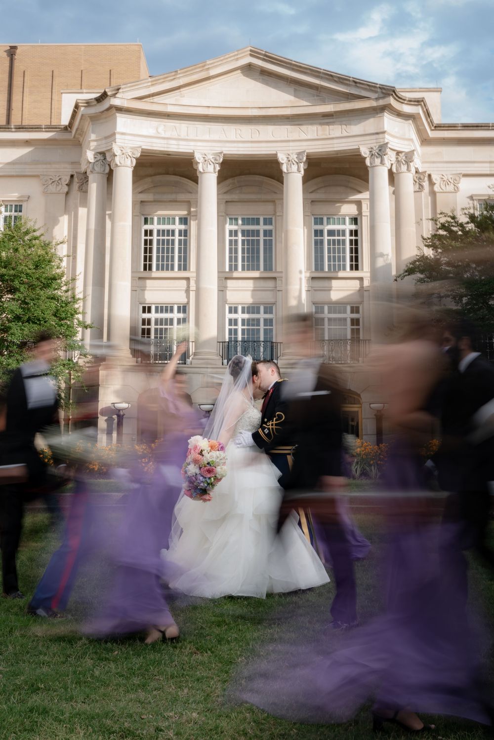 Bride and groom pose outside elegant columned building while wedding party moves in blur around them.