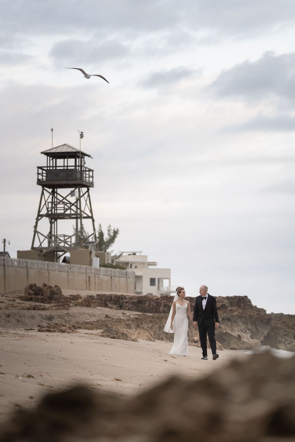 Bride and groom walking hand in hand on a sandy beach with a lifeguard tower and seagull in the background.