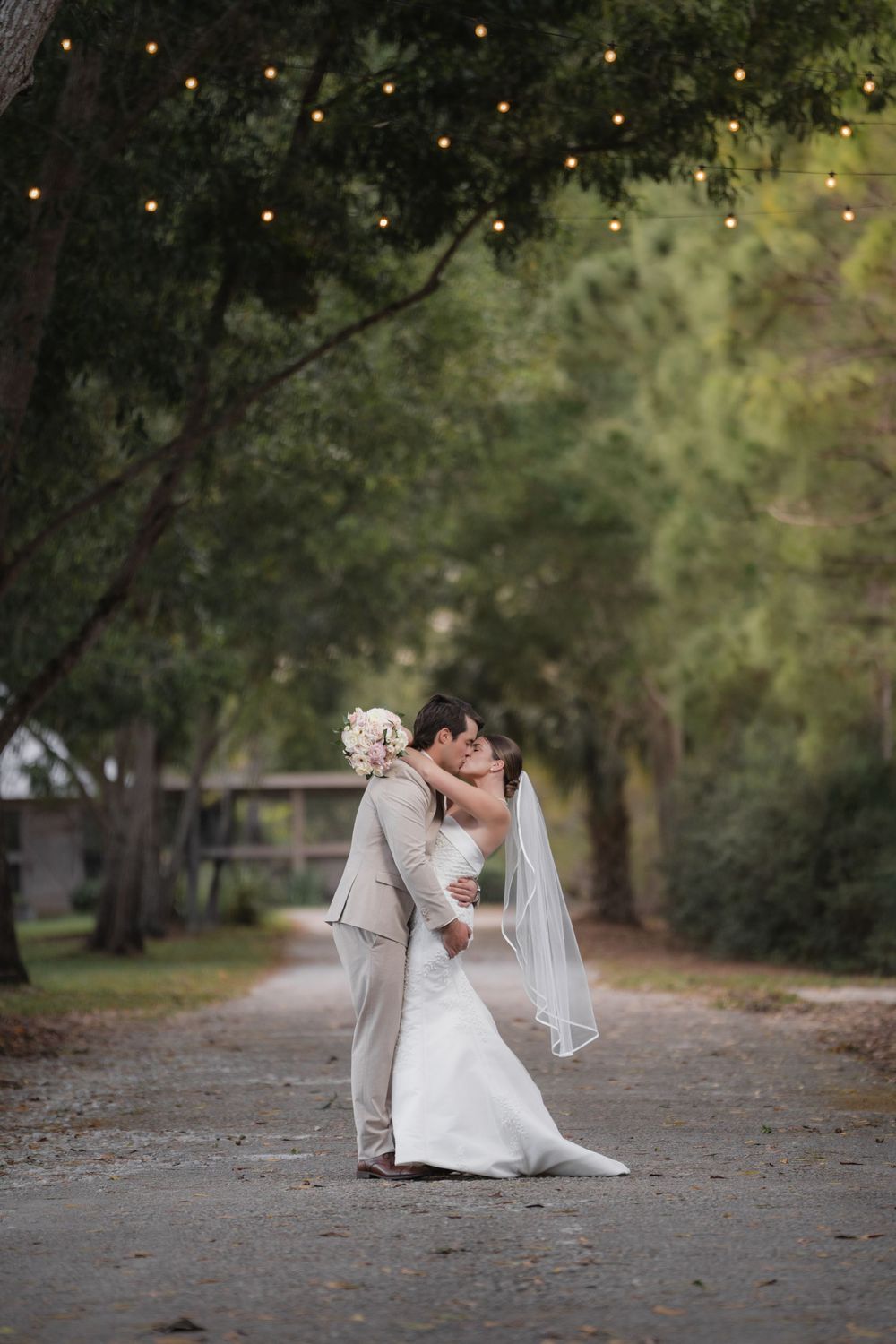 Bride and groom share a romantic kiss on a tree-lined path adorned with string lights at their outdoor wedding.