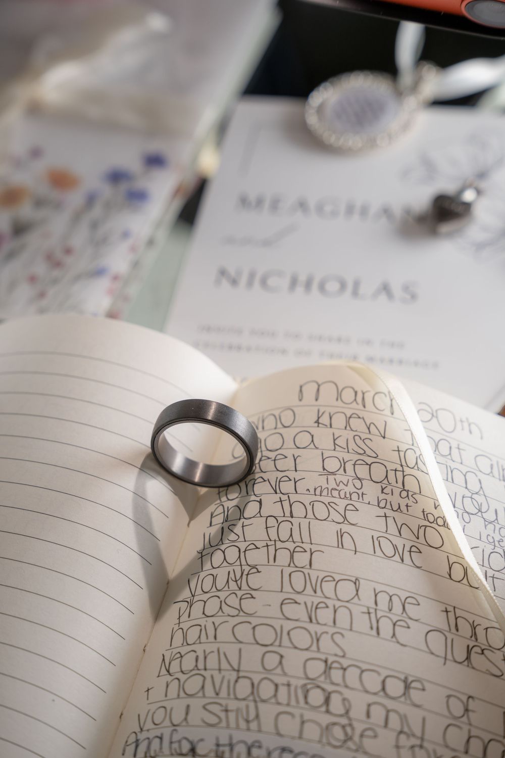 A silver wedding band rests on an open journal with handwritten vows, beside a Meaghan and Nicholas wedding invitation.