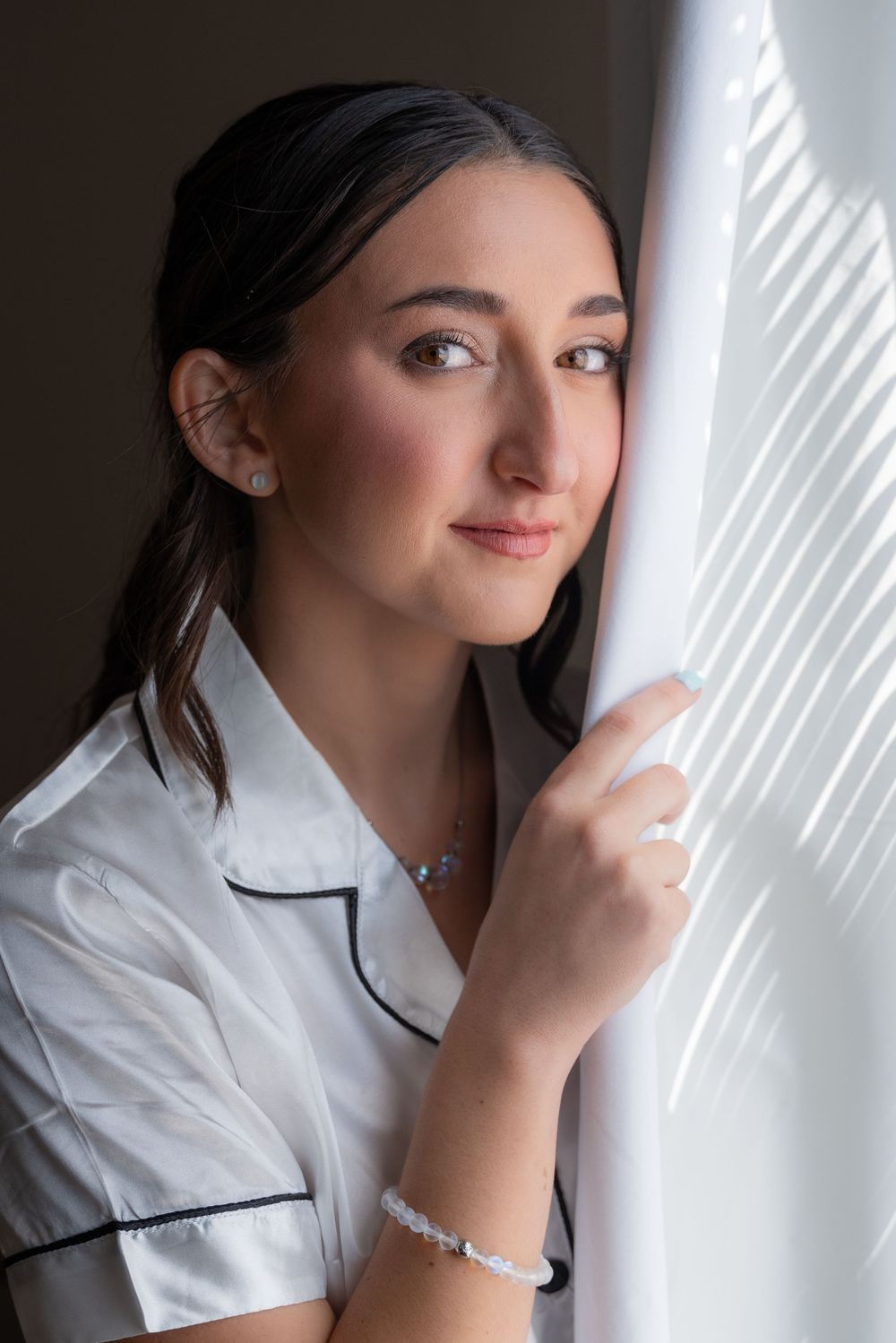 Young woman in white pajamas leans against window curtain, wearing silver jewelry, soft natural light.