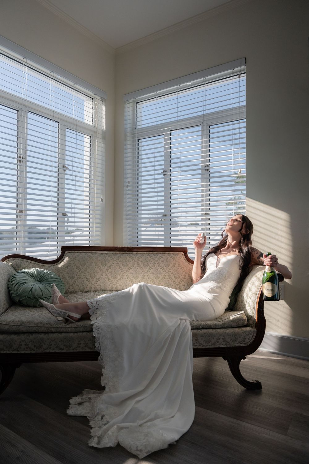 Bride in white gown lounging on vintage chaise with champagne bottle, bathed in natural light from white shuttered windows.