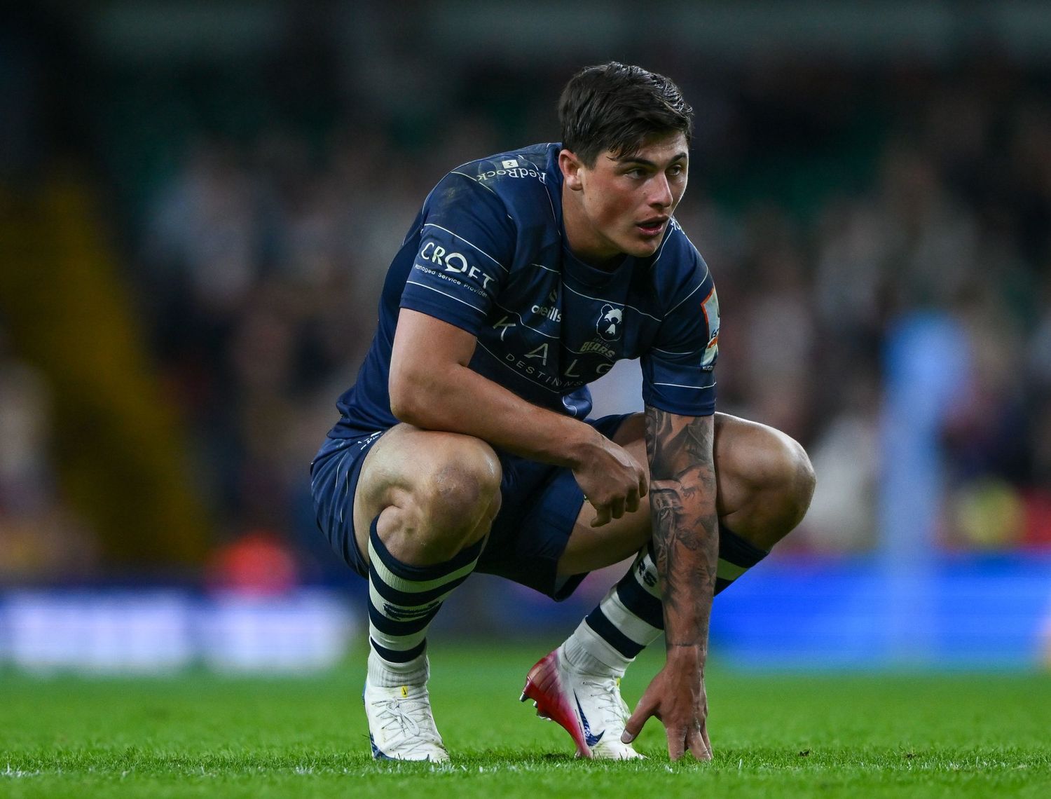 Rugby player in dark blue jersey with KAELI sponsor logo stands on field during a match.