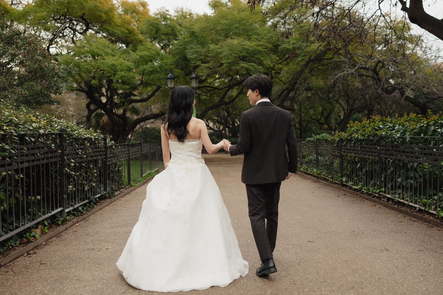 A couple in wedding attire walks hand in hand down a tree-lined garden path surrounded by lush greenery.