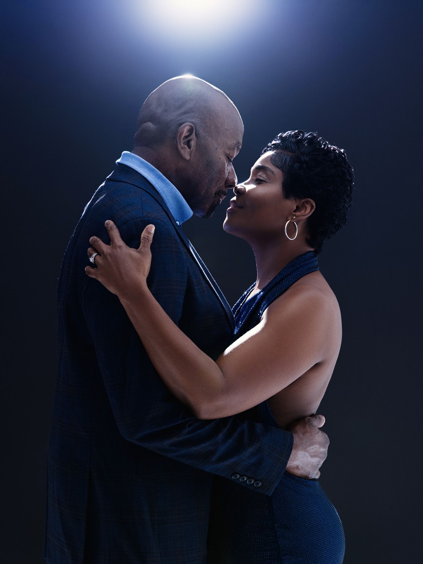 A couple shares an intimate dance moment in a dimly lit setting wearing formal attire against a dark background.