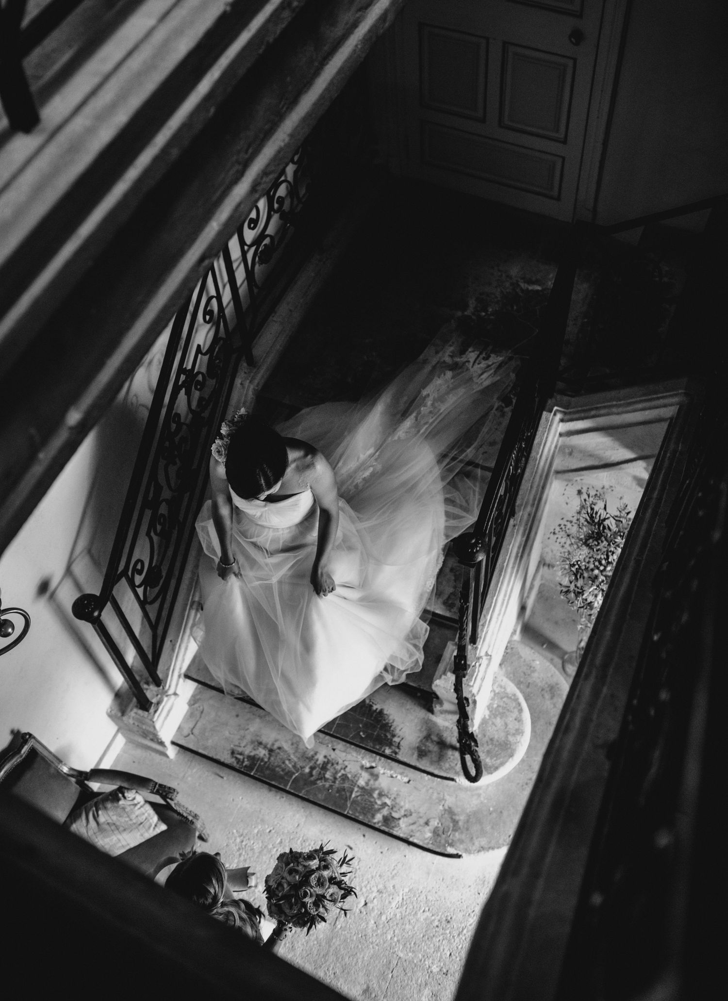 Bride walking down a staircase inside a French château with flowing veil, captured from above in a timeless black and white editorial style