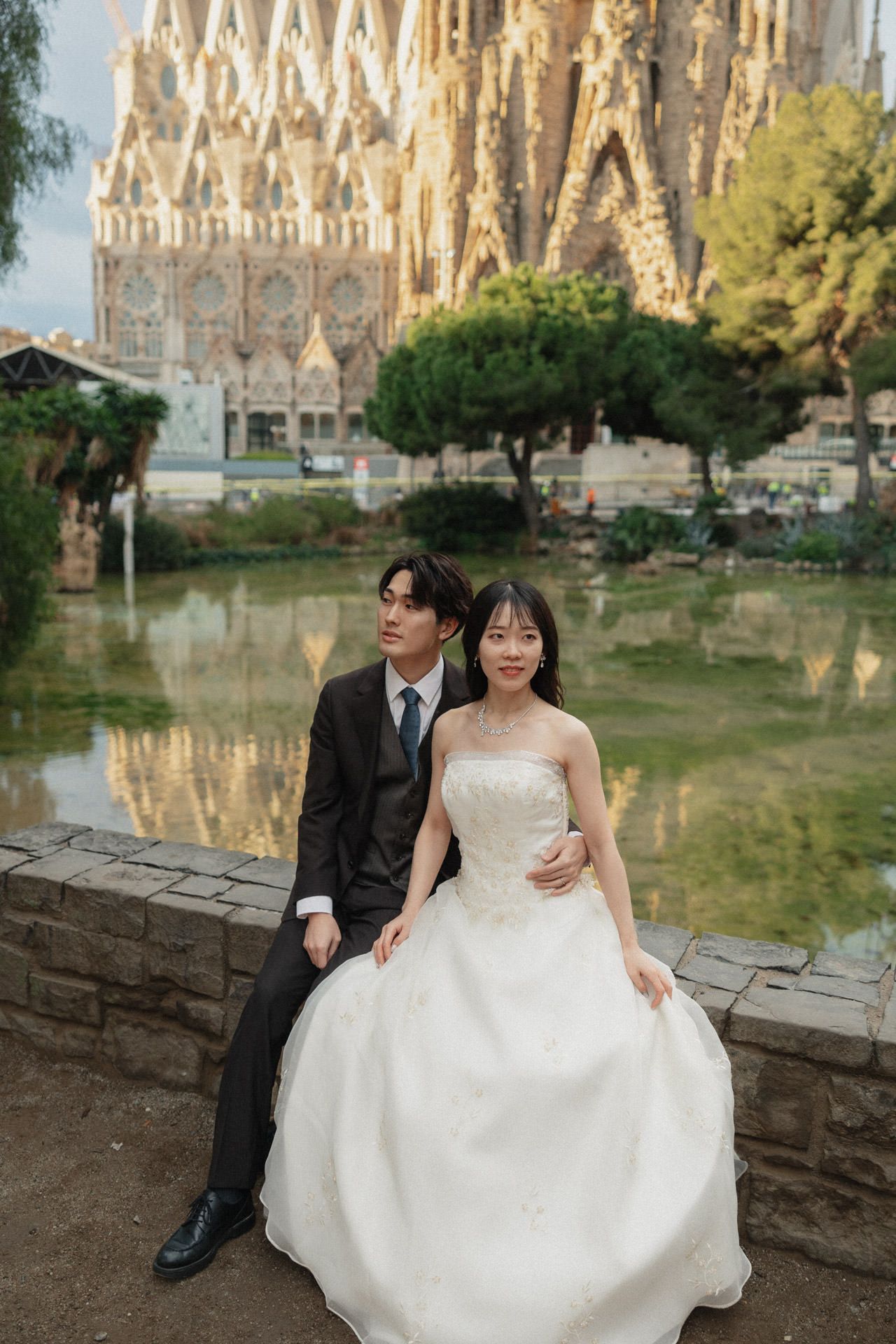 A couple in wedding attire sits on a stone wall with an ornate Sagrada Família in Barcelona and reflecting pool in the background.
