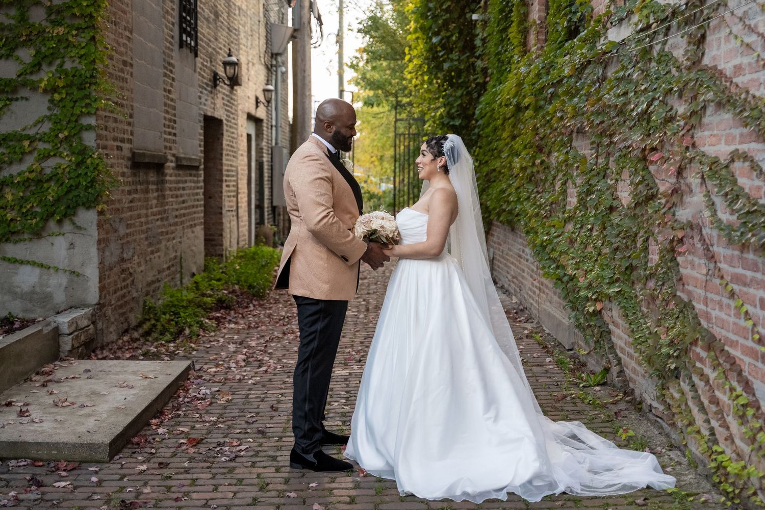 A couple in wedding attire walks along a charming stone pathway surrounded by lush greenery and brick walls.