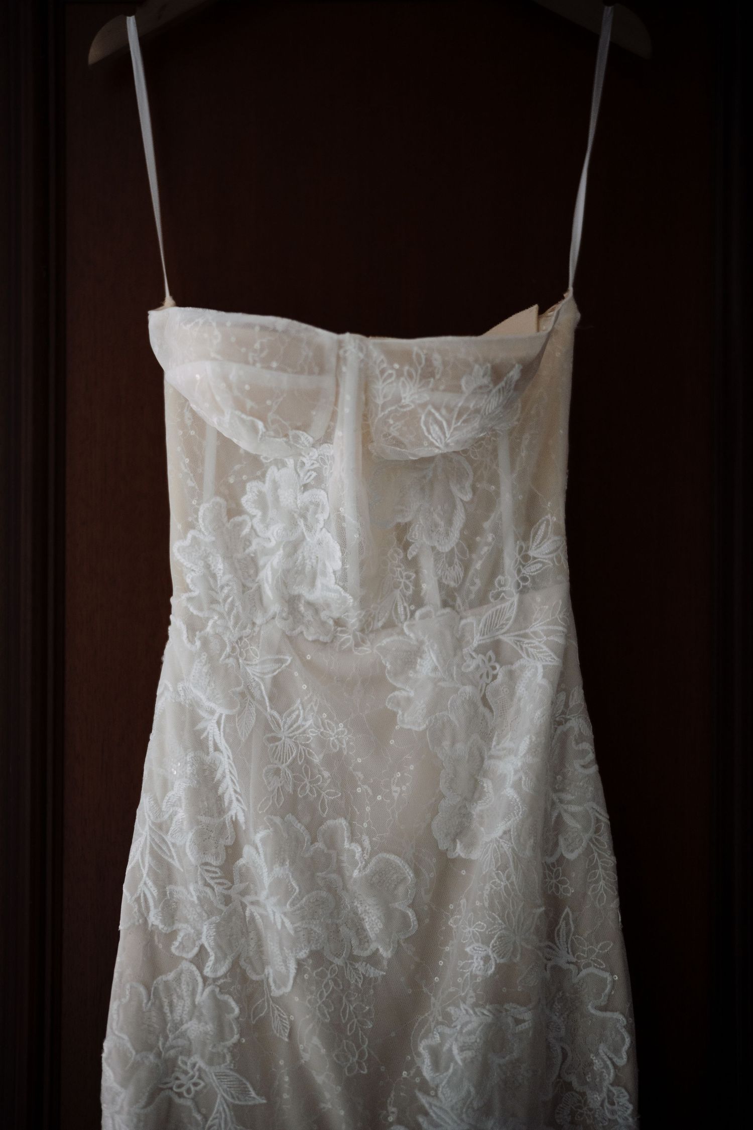 Elegant white lace wedding dress with spaghetti straps and floral embroidery hanging on a dark wooden door.