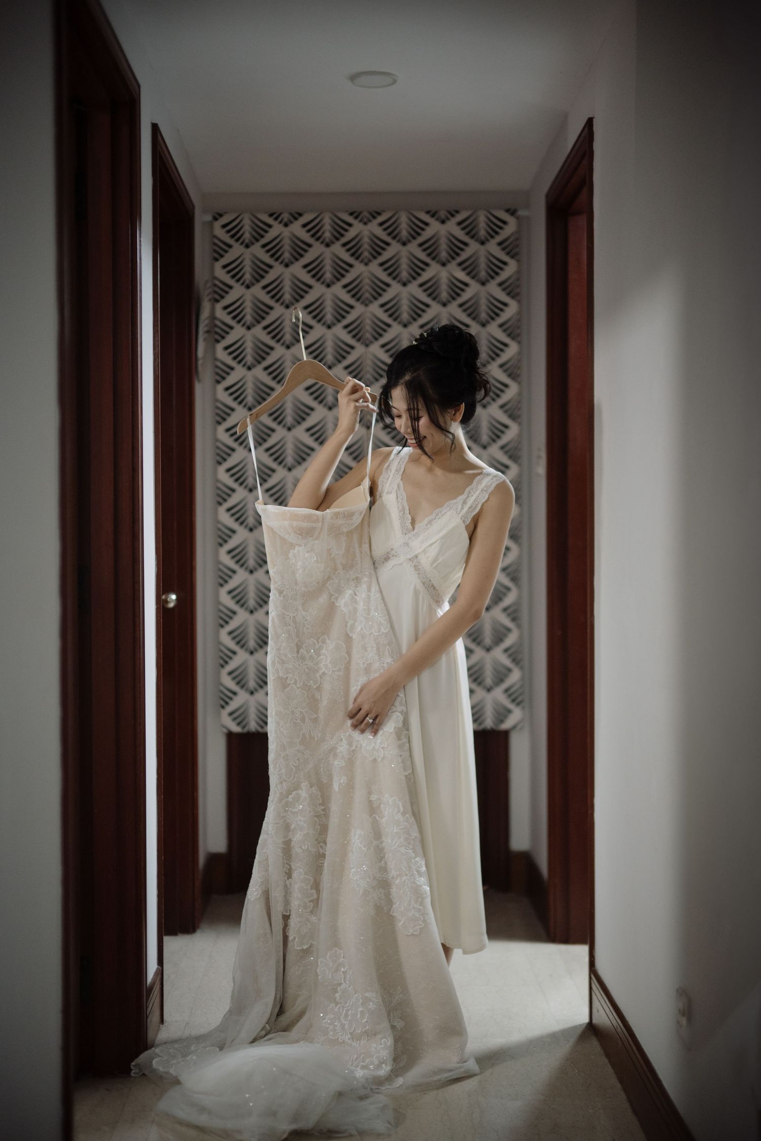 Bride in a white robe holding her lace wedding dress on a hanger in a hotel corridor.