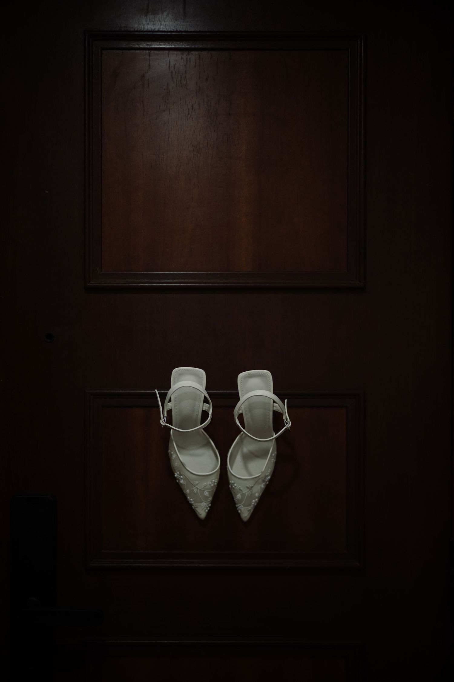 White pearl-embellished bridal heels hanging on a dark wooden door panel before the wedding ceremony.
