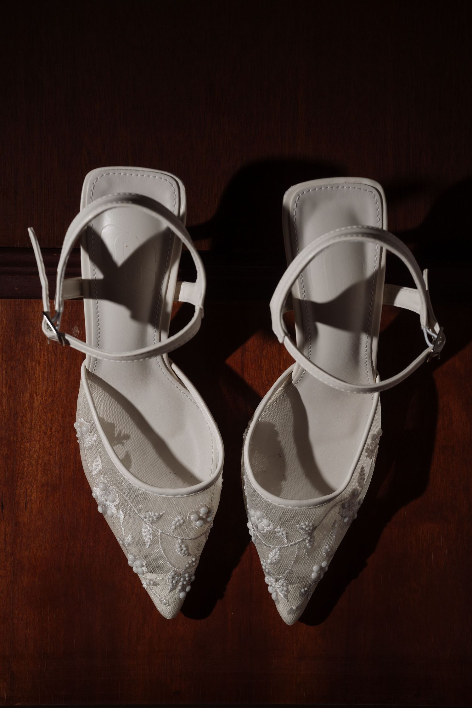 White embroidered bridal heels with ankle straps and floral detailing on a dark wooden surface.