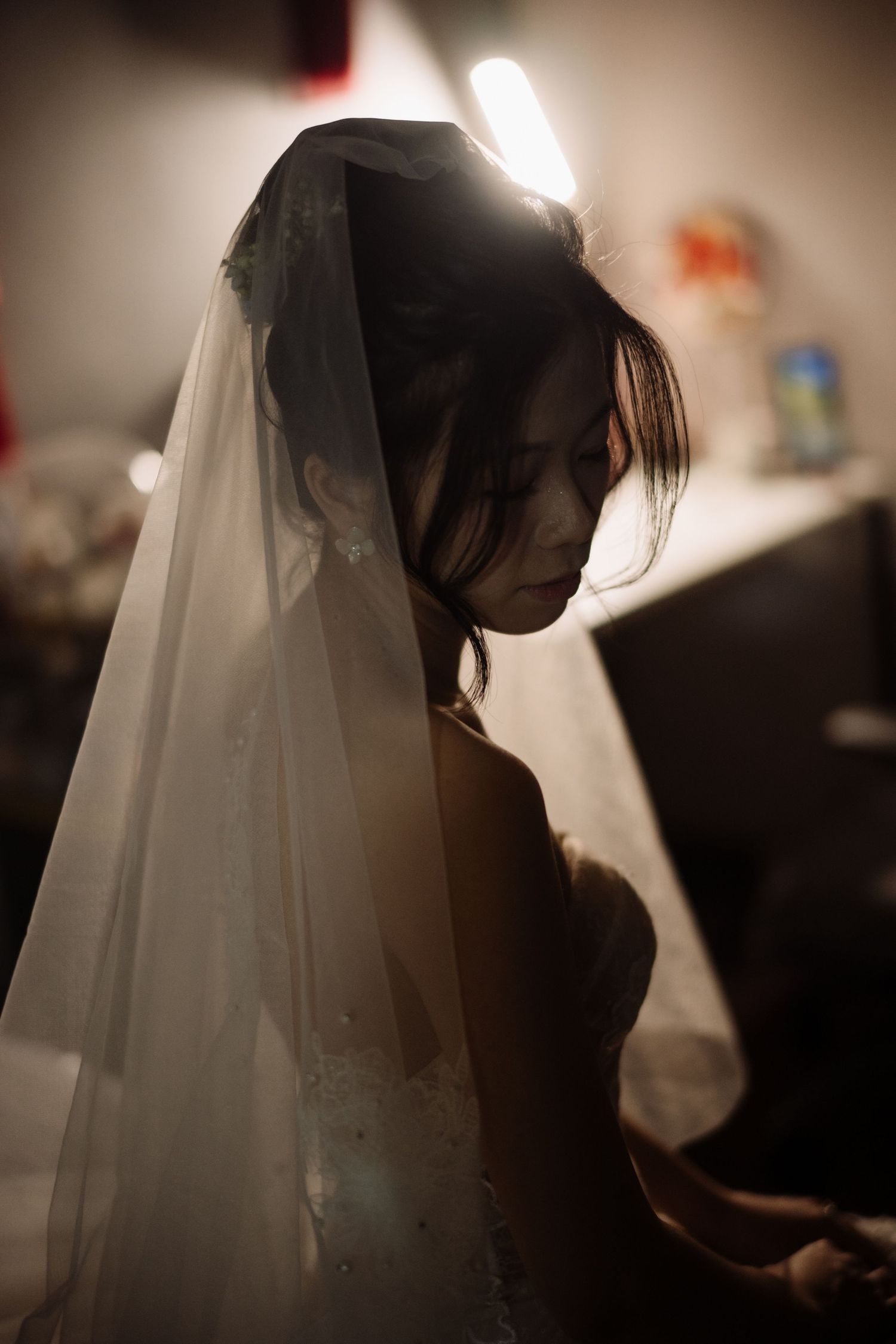 Bride wearing a delicate white veil with her head bowed in a softly lit room before the wedding.