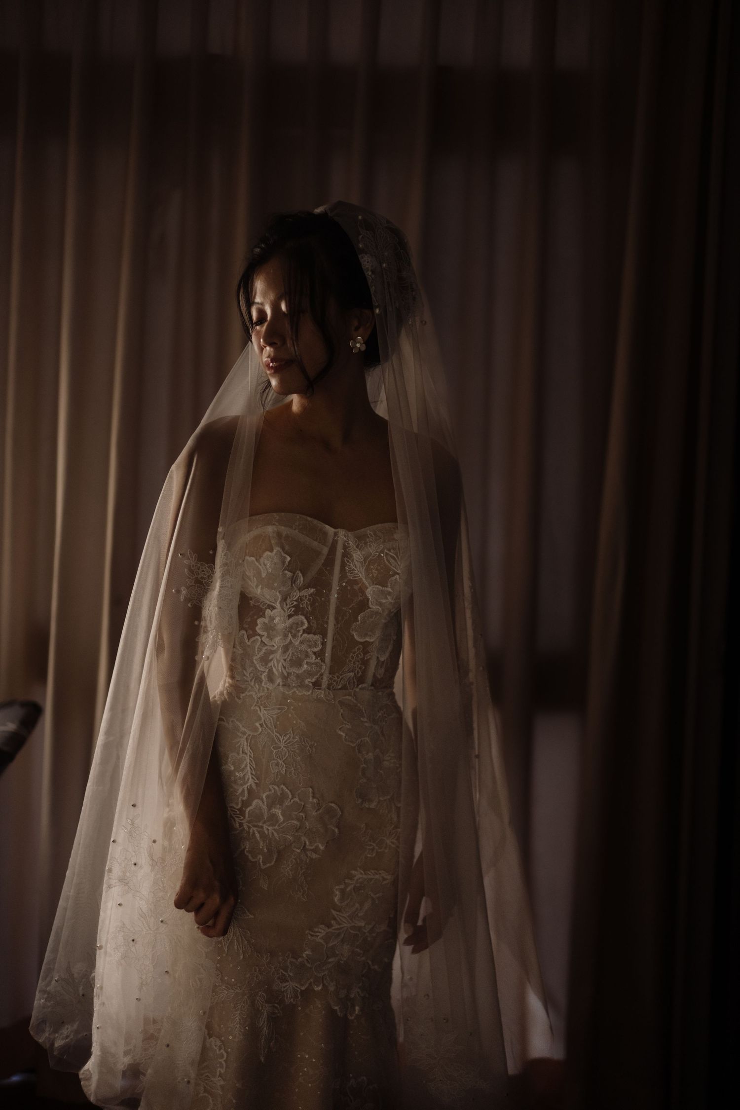 Elegant bride in a fitted lace wedding gown and long veil standing by a curtained window in dim light.