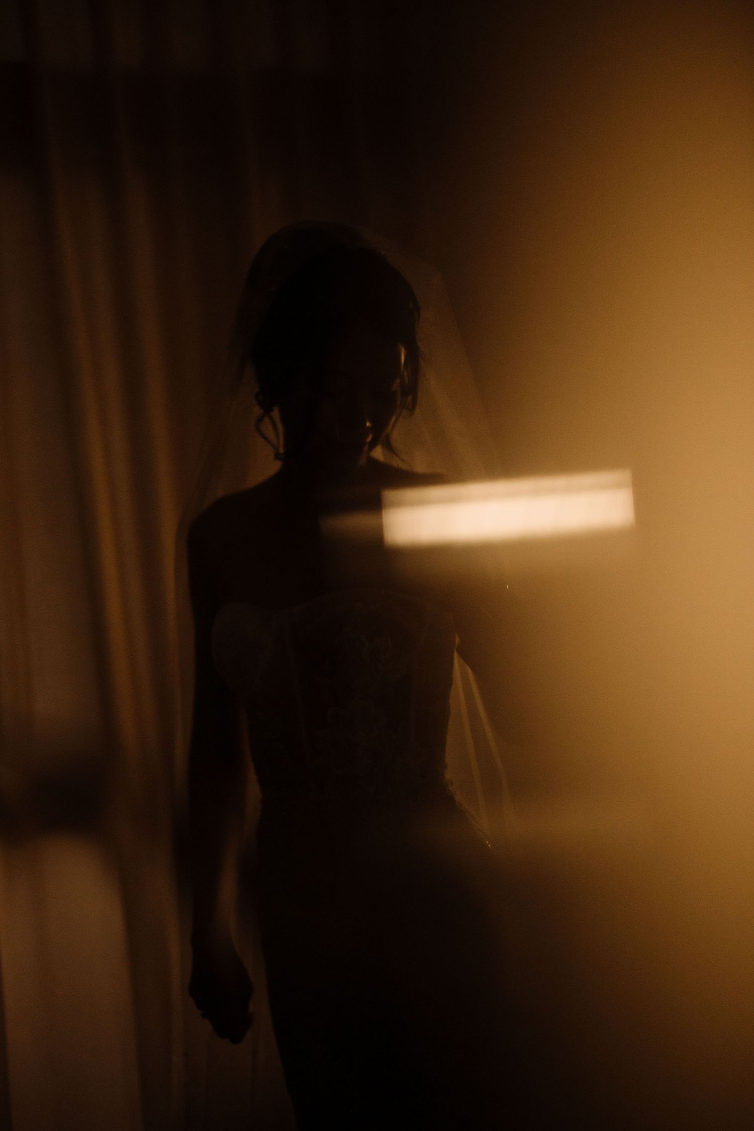 Dramatic silhouette of a bride in a wedding dress backlit by warm golden light through a curtain.
