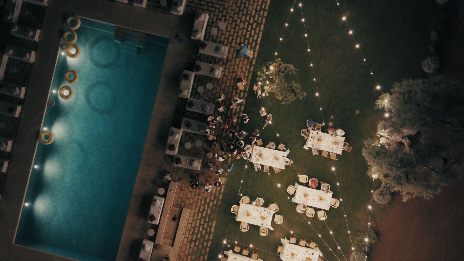 Aerial night view of an outdoor wedding reception with string lights, white tables, and a glowing pool.
