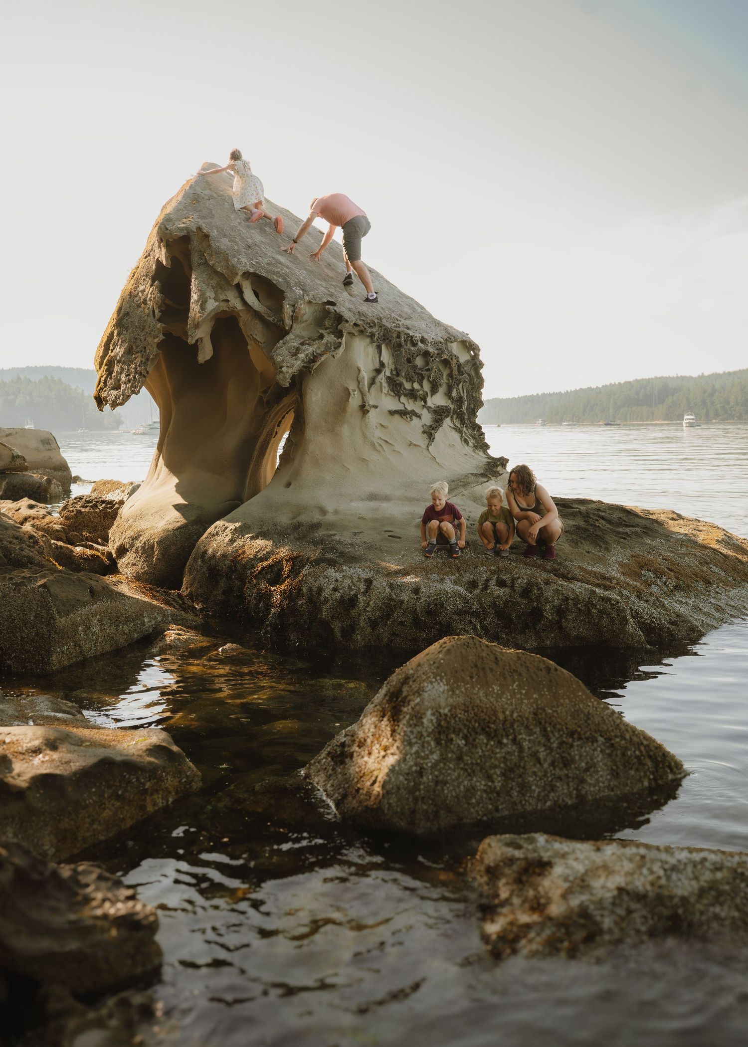 People climbing and sitting on a large sculpted rock formation by the ocean shore with forested hills in the background.