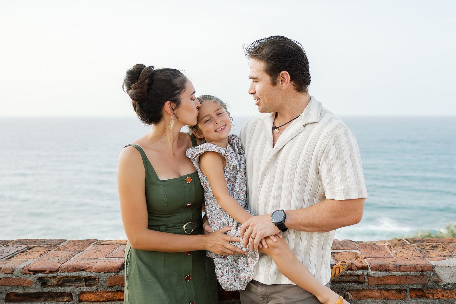 A family of three, a mother, father, and young child, standing together outdoors with the ocean in the background.