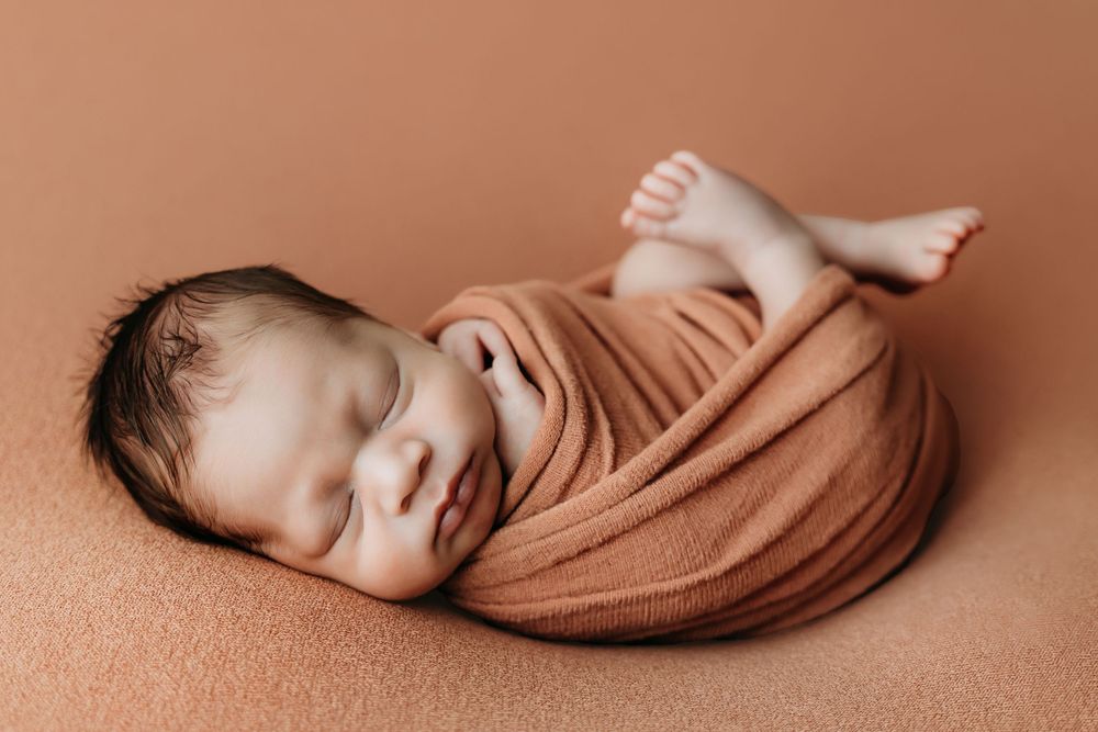 Newborn baby swaddled in a warm terracotta wrap, peacefully sleeping on a matching brown backdrop.
