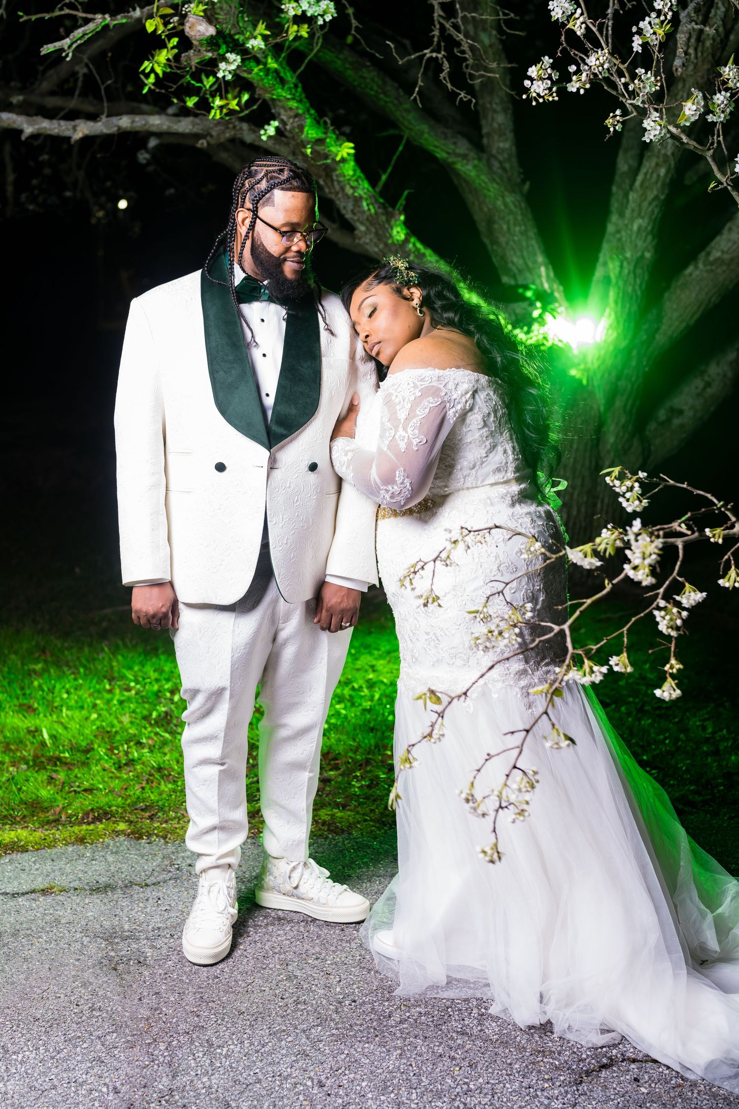 A romantic wedding couple poses in white attire under illuminated trees at night during their outdoor photo session.