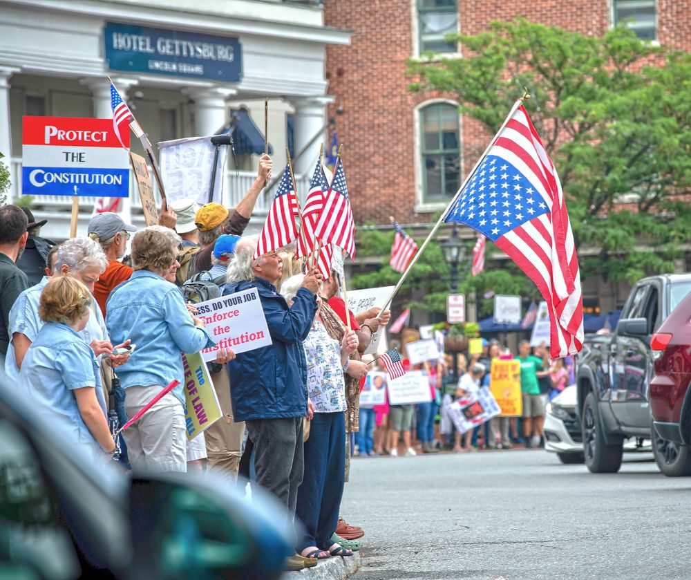 Crowd gathered along a brick building sidewalk waving American flags during a patriotic event.