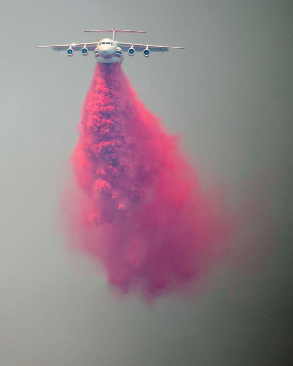 Large aircraft drops vibrant red fire retardant in a dramatic cloud formation against grey sky.