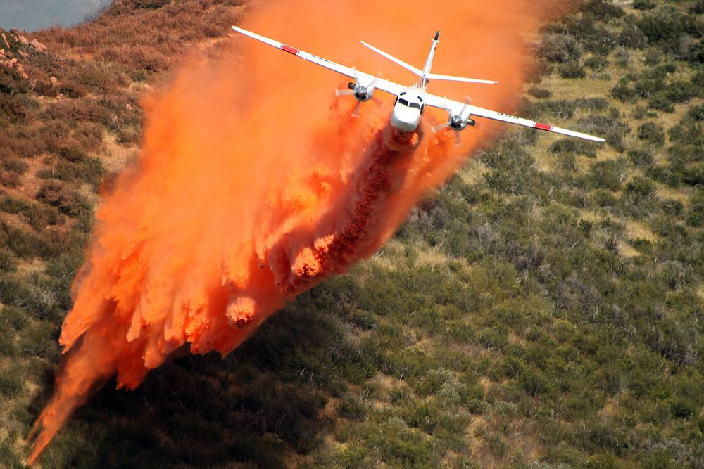 Aerial firefighting plane dropping orange fire retardant over brush during wildfire response.