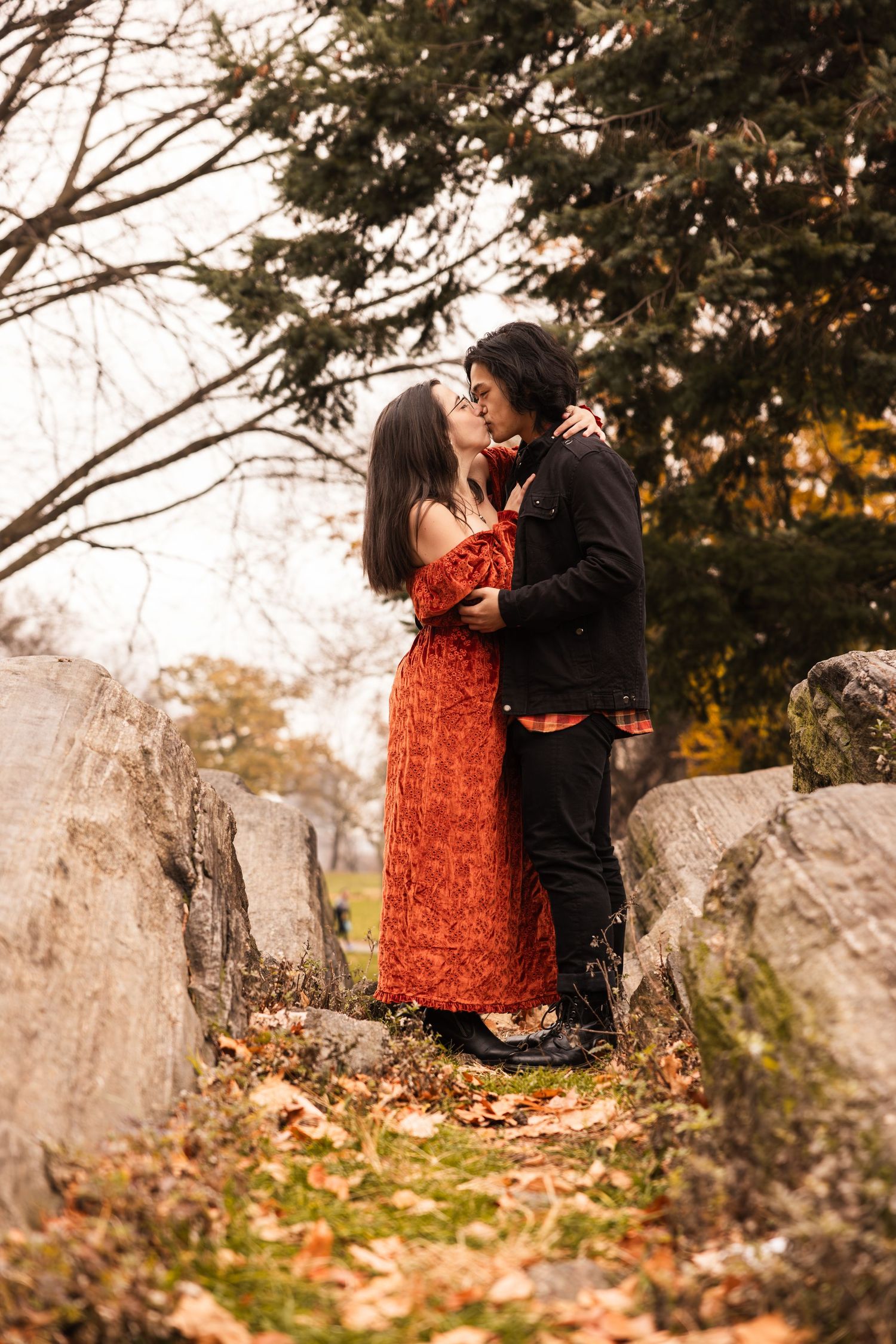 A romantic autumn scene shows a couple embracing on stone steps surrounded by fallen leaves and evergreen trees.
