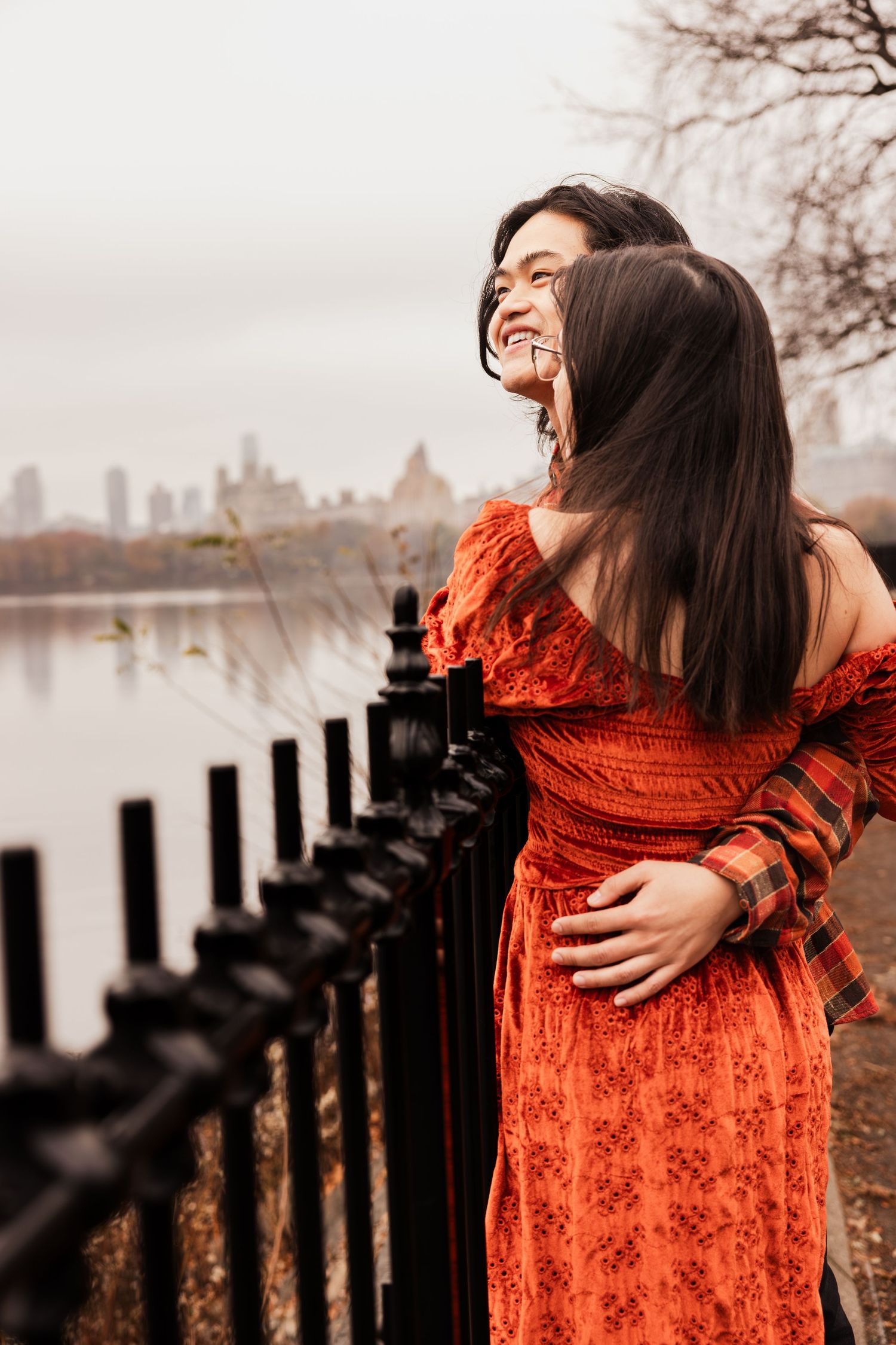 A person in a flowing orange lace dress poses dramatically against a wooden fence by a misty waterfront.