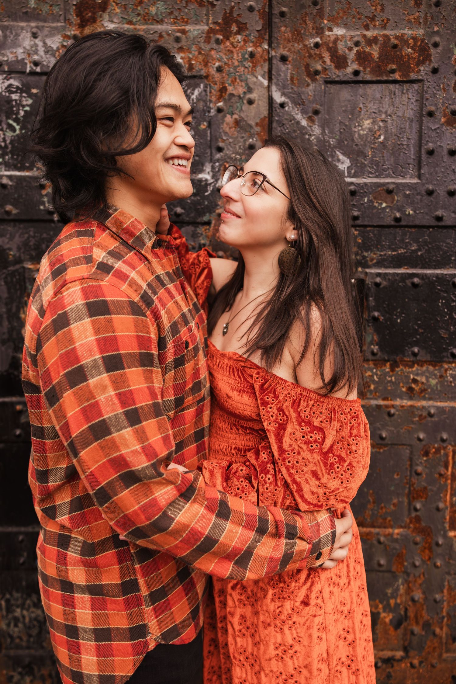 A couple in coordinating orange outfits share a romantic moment against a rustic metal door background.