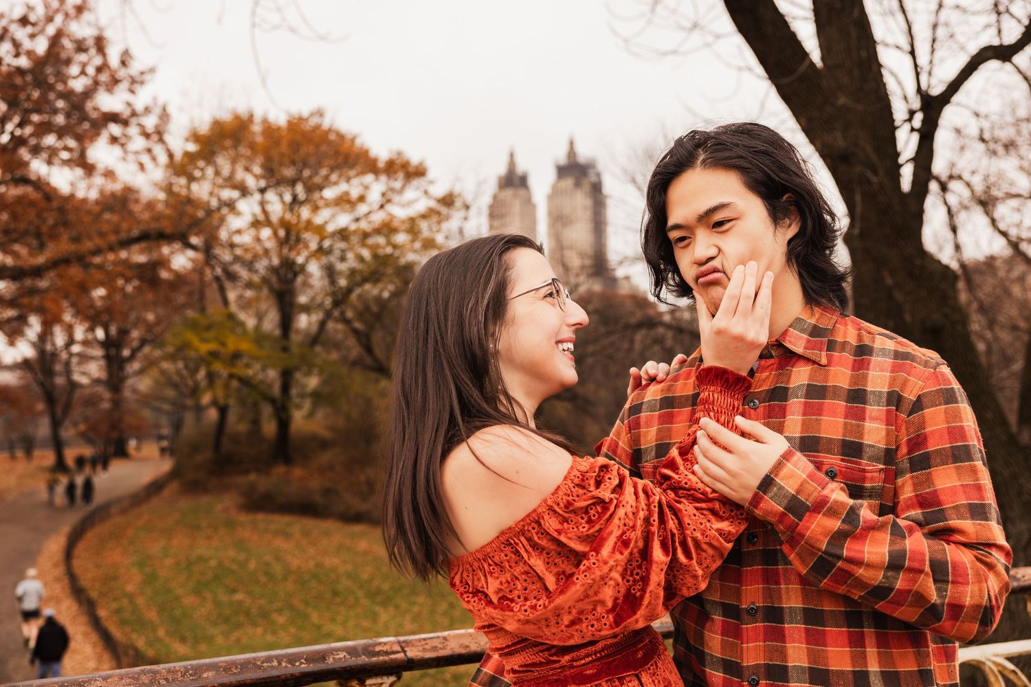 A couple in autumn attire embraces and laughs together in a park with fall foliage in the background.