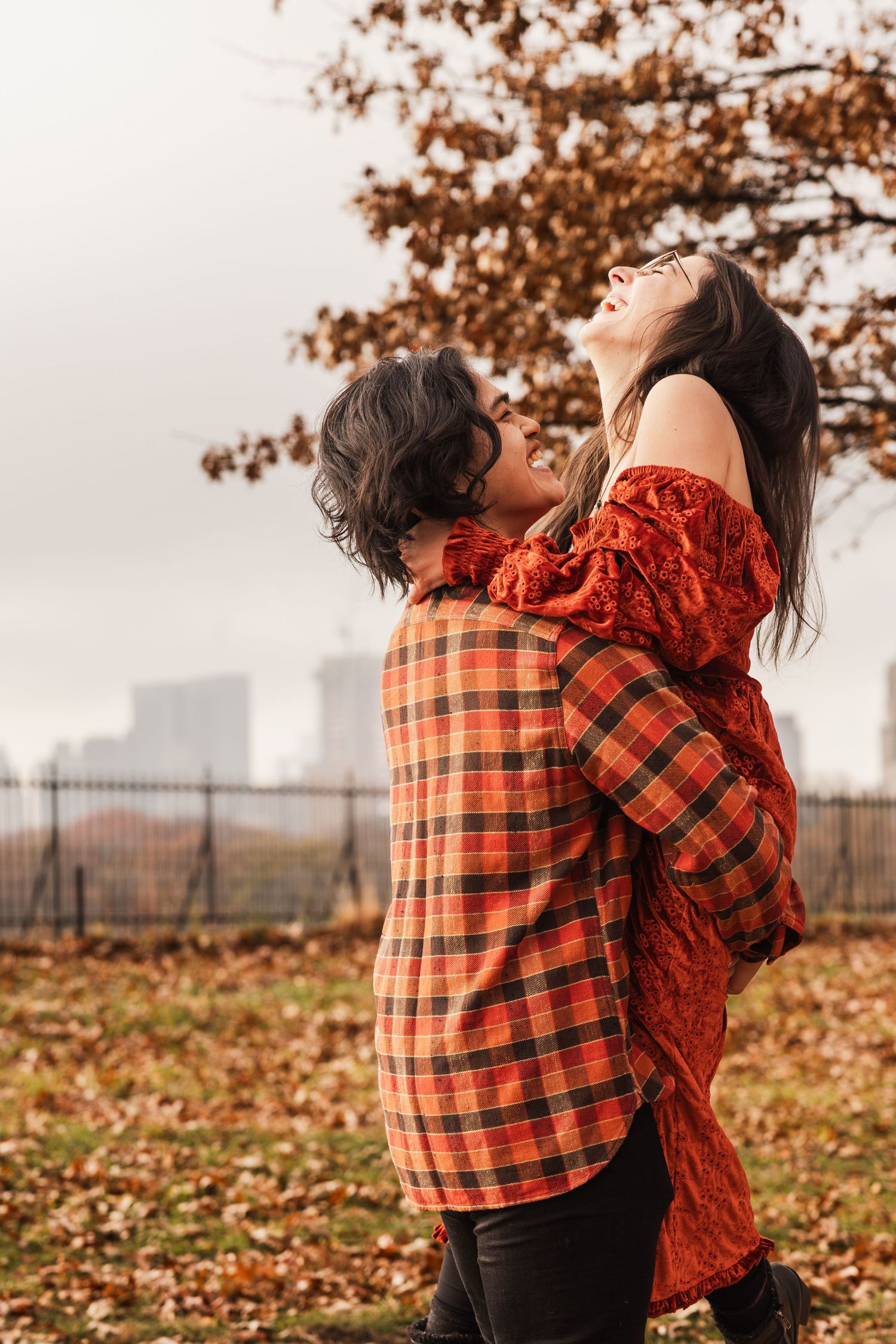 Couple embraces in autumn park with orange plaid shirt and red dress against fall foliage and city skyline background.