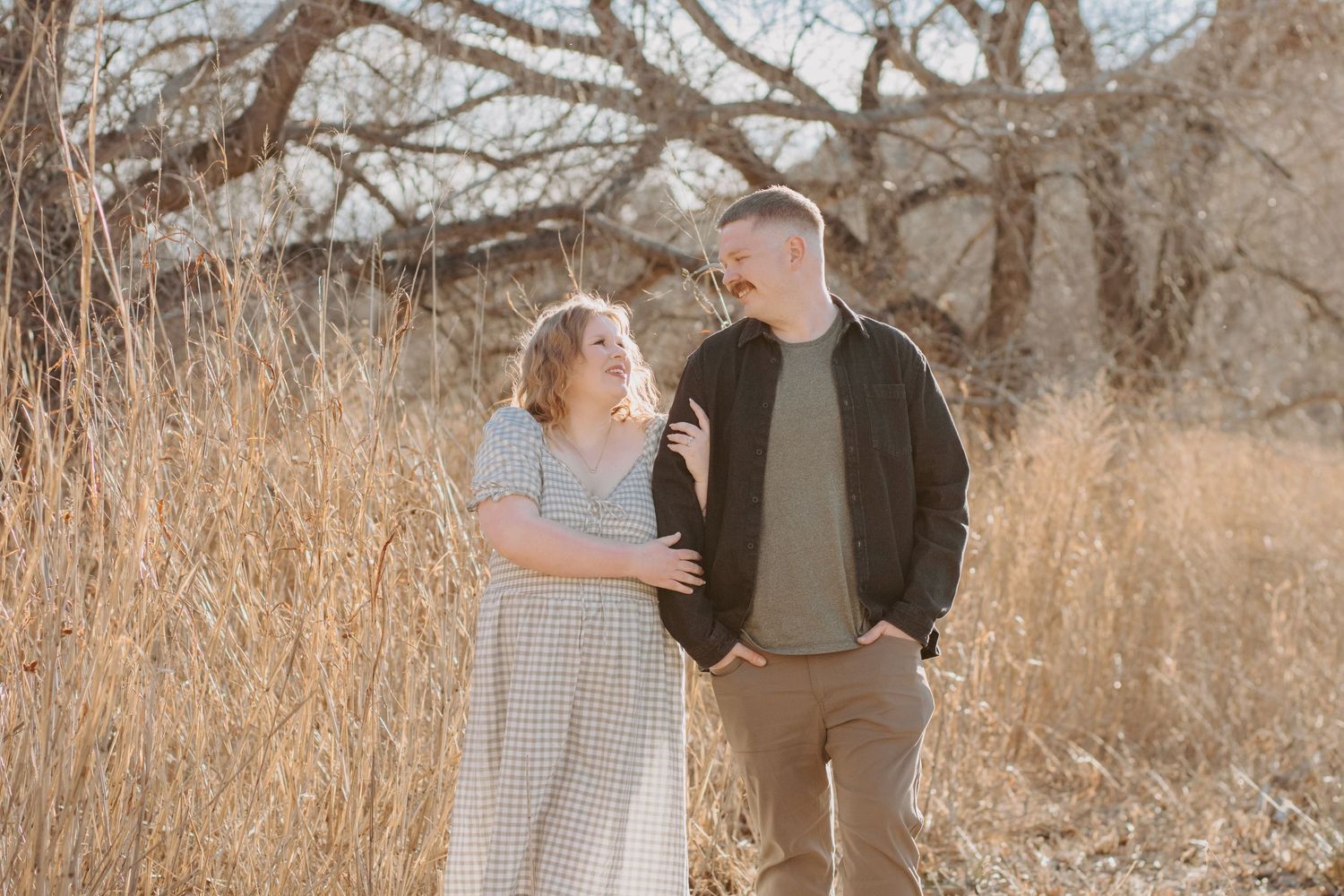 Couple smiling at each other in a field of tall golden grass with bare trees in the background.