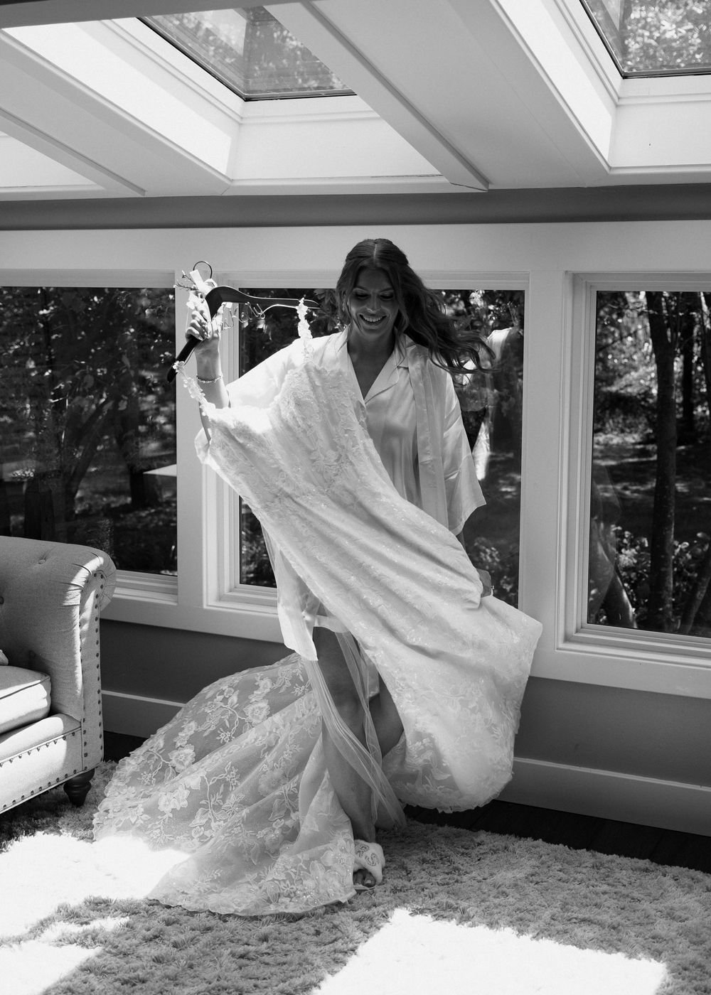 Black and white photo of a joyful bride twirling her lace wedding gown in a sunlit room with large windows.