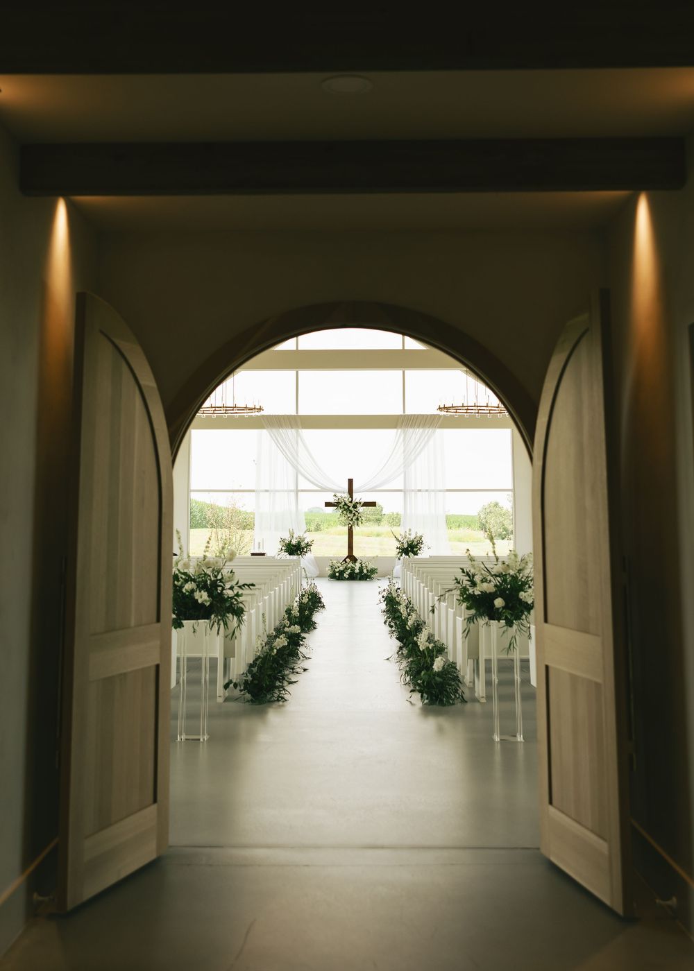 Elegant wedding chapel interior with arched doors, white pews, greenery aisle decor, and a cross altar bathed in natural light.