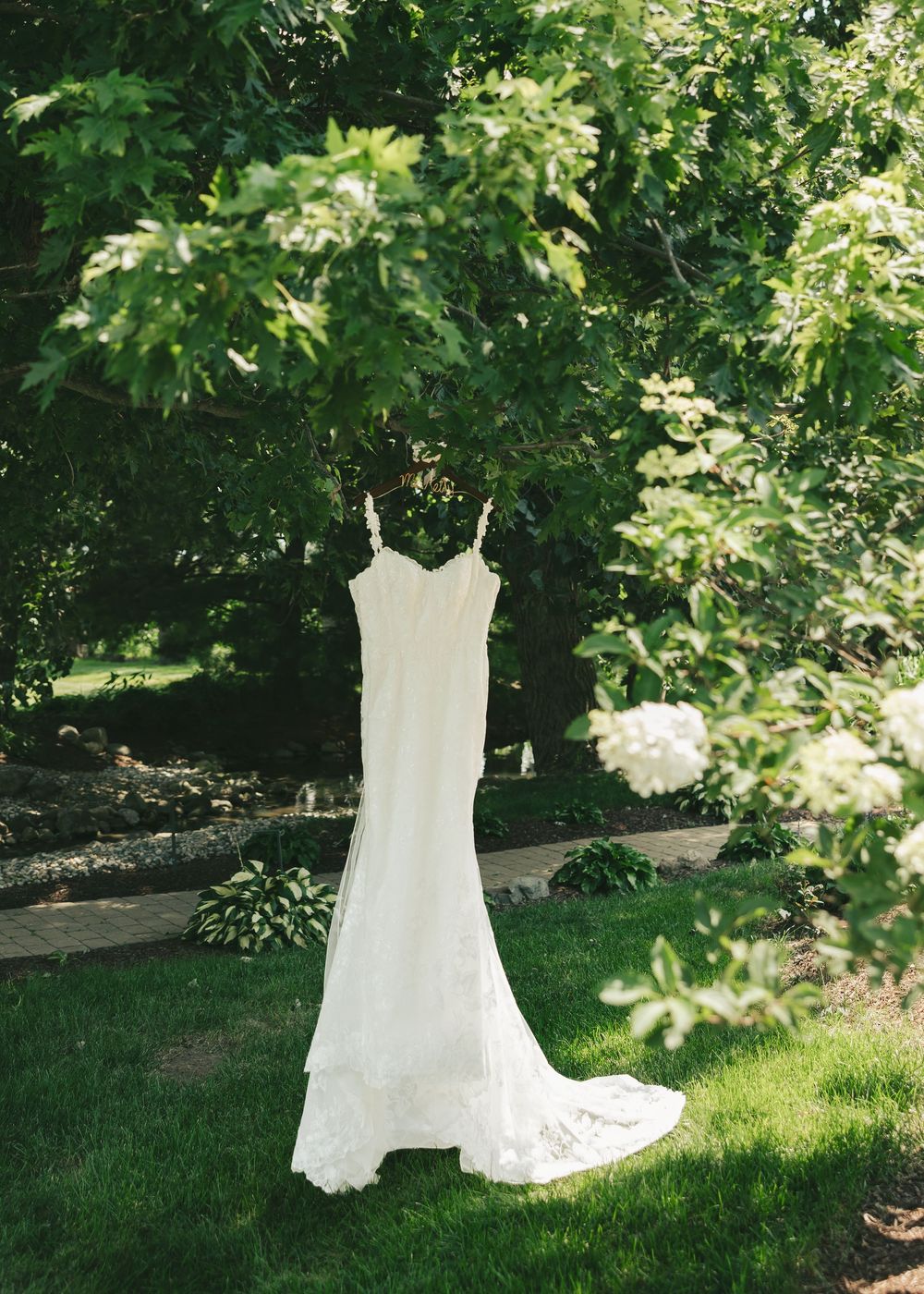 White lace wedding gown with spaghetti straps and train hanging outdoors among lush green trees and hydrangeas.